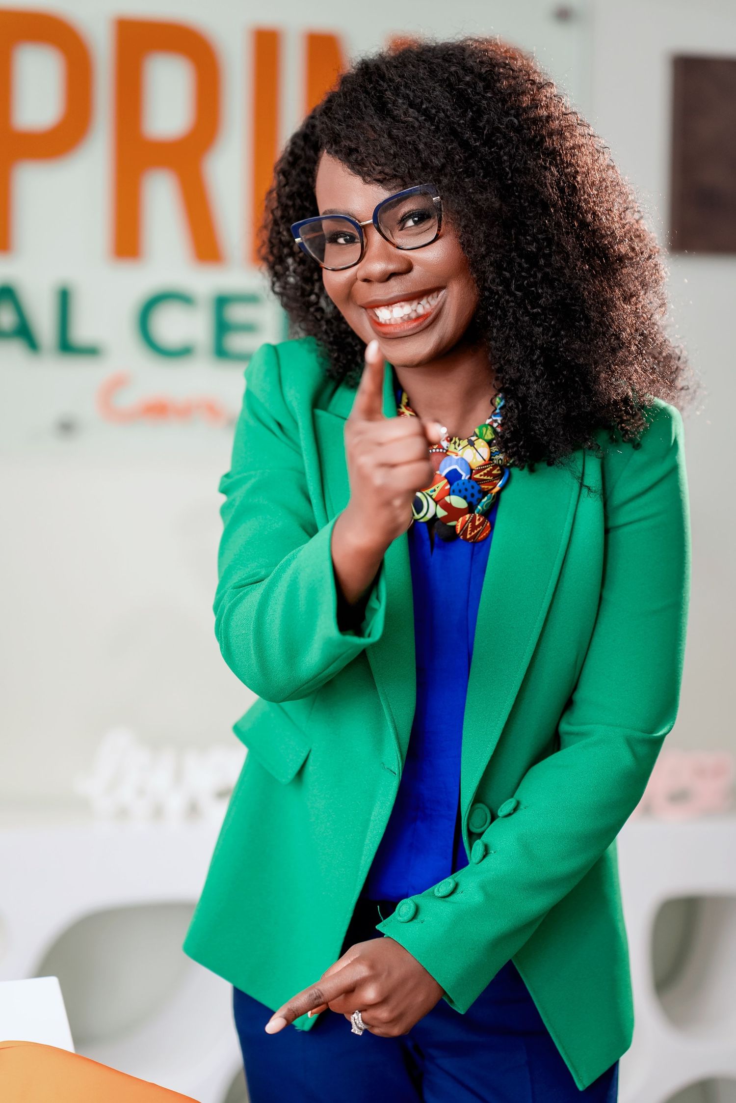 Professional wearing a vibrant green blazer and blue dress with glasses giving thumbs up in an office setting.