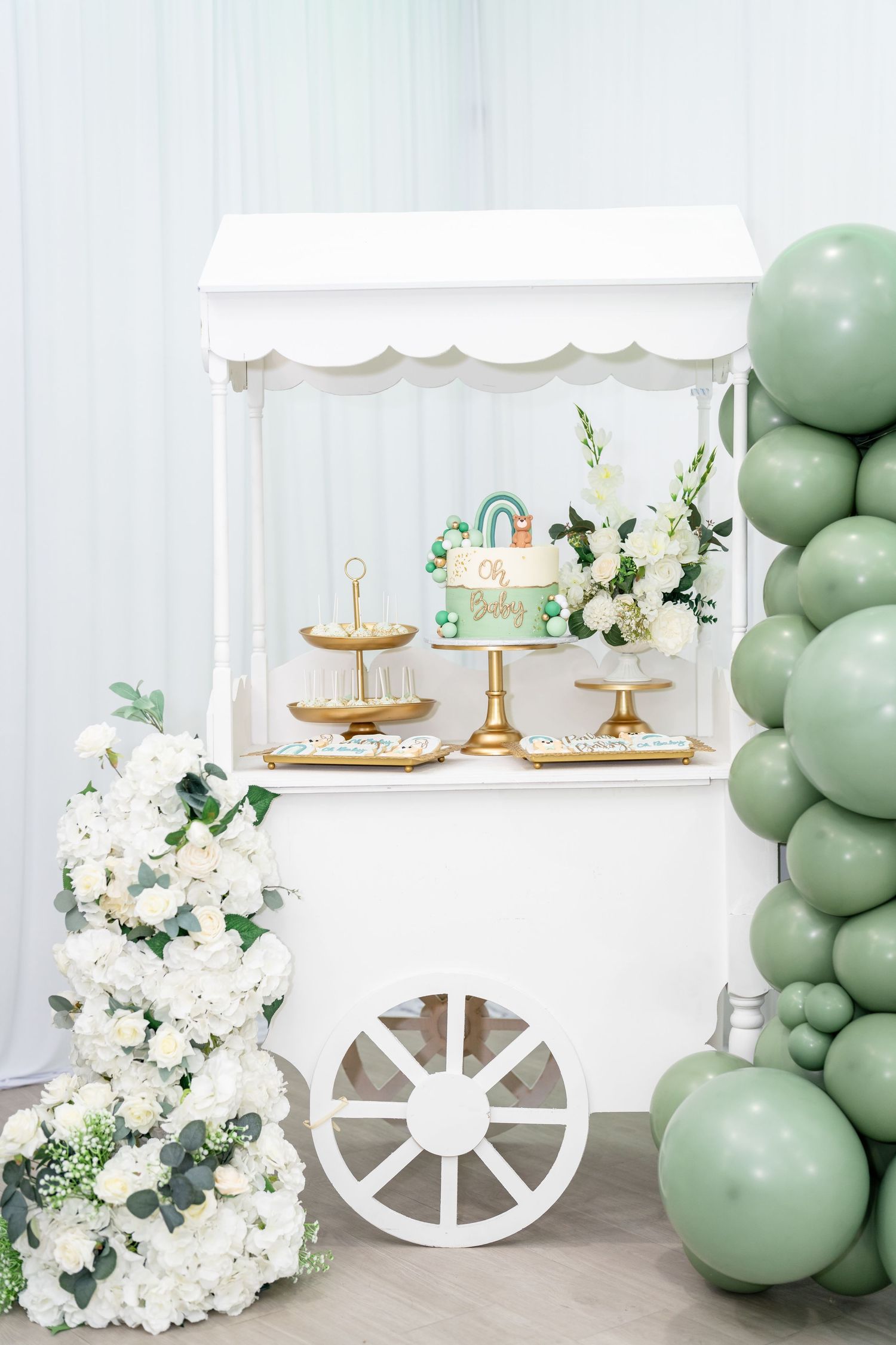 White dessert cart decorated with sage green balloons, white flowers and eucalyptus for an elegant party setup.