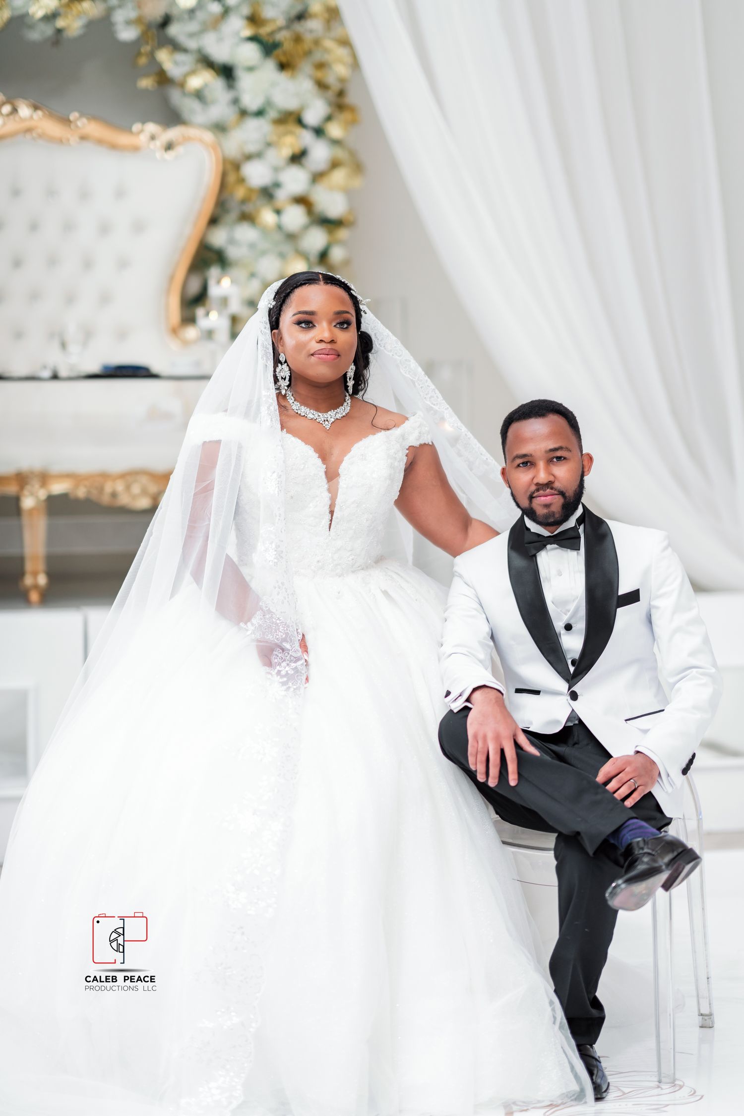 Elegant wedding photoshoot featuring bride in white gown and groom in black tuxedo against white decor and floral backdrop.