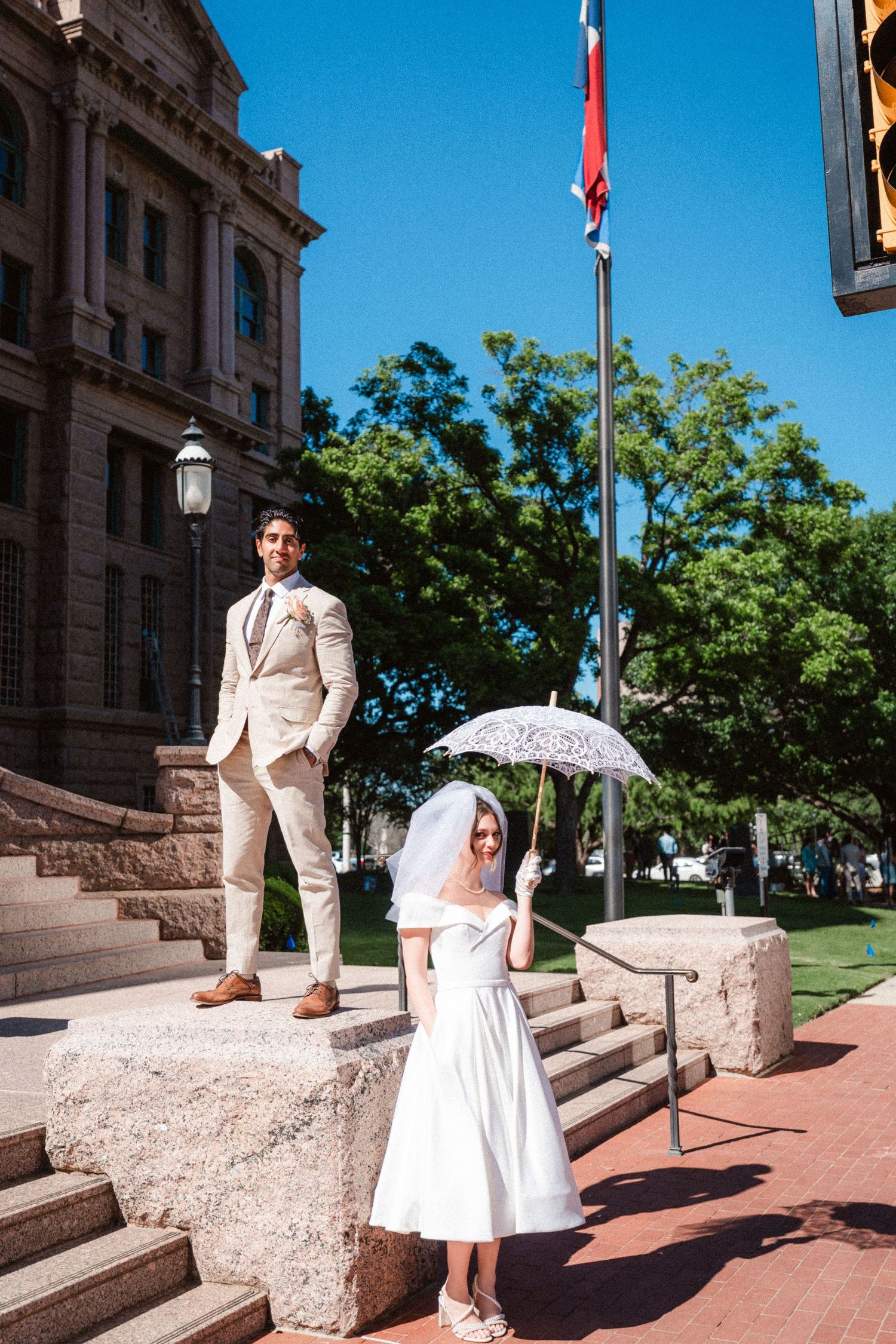 A bride and groom pose on stone steps outside a government building with flags.