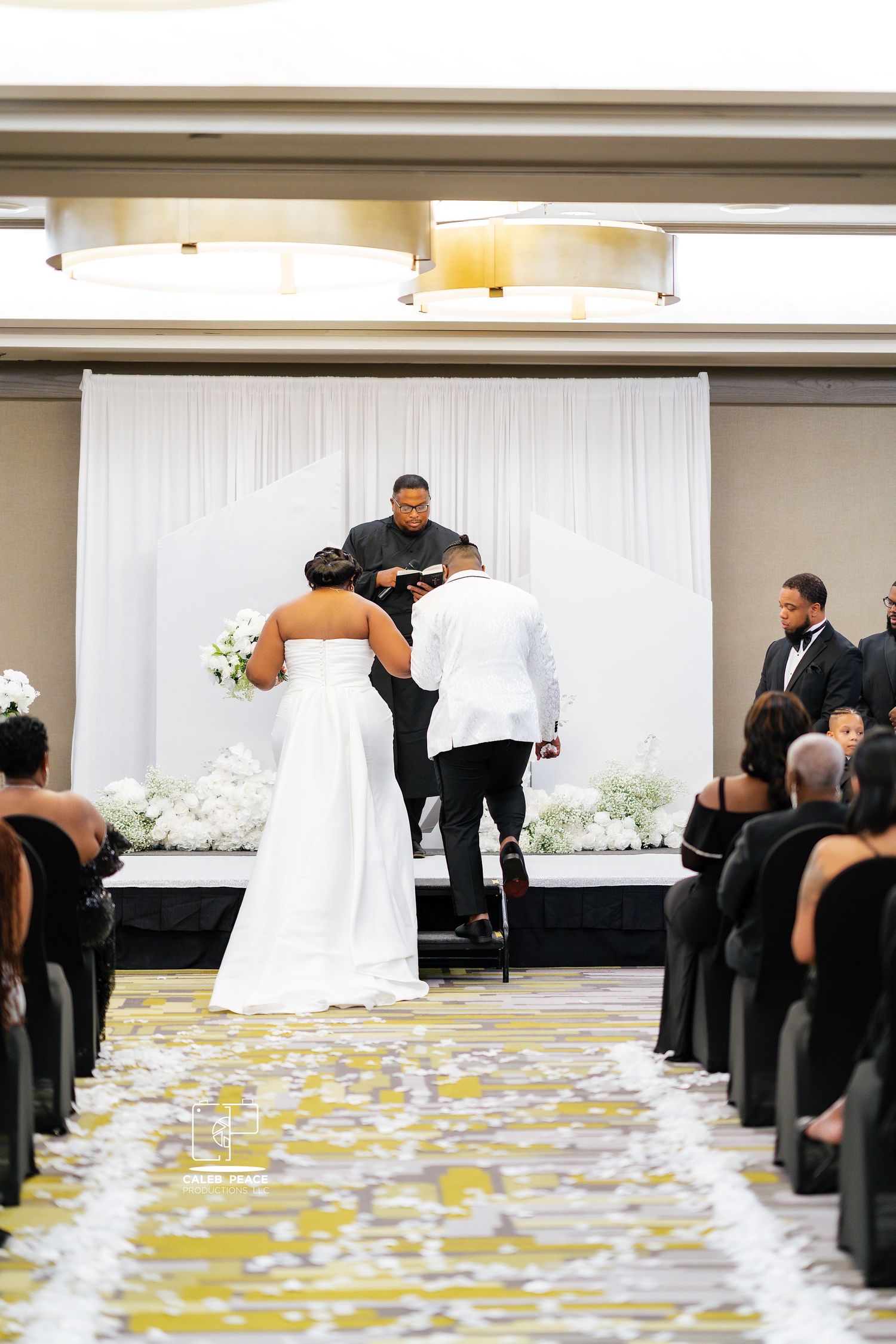 Wedding guests gather around a white-draped table with scattered yellow flower petals in an elegant reception hall.