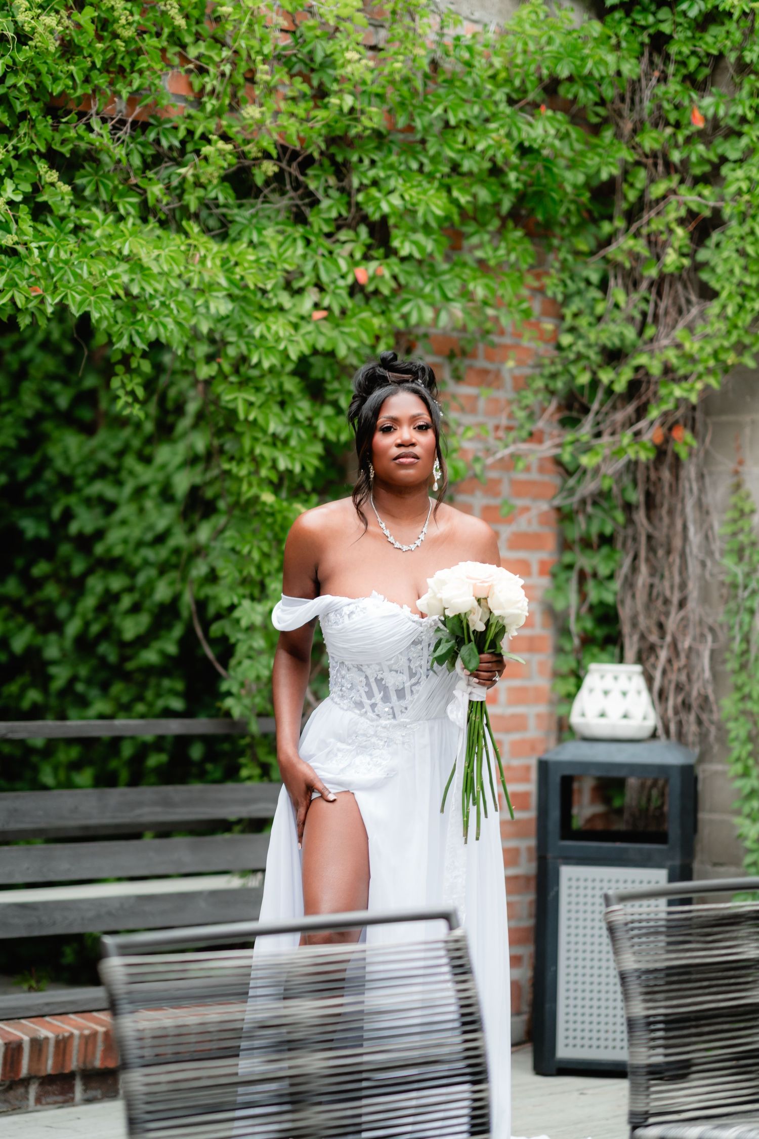 A bride in a white lace gown poses elegantly with a bouquet against a backdrop of green foliage and brick walls.