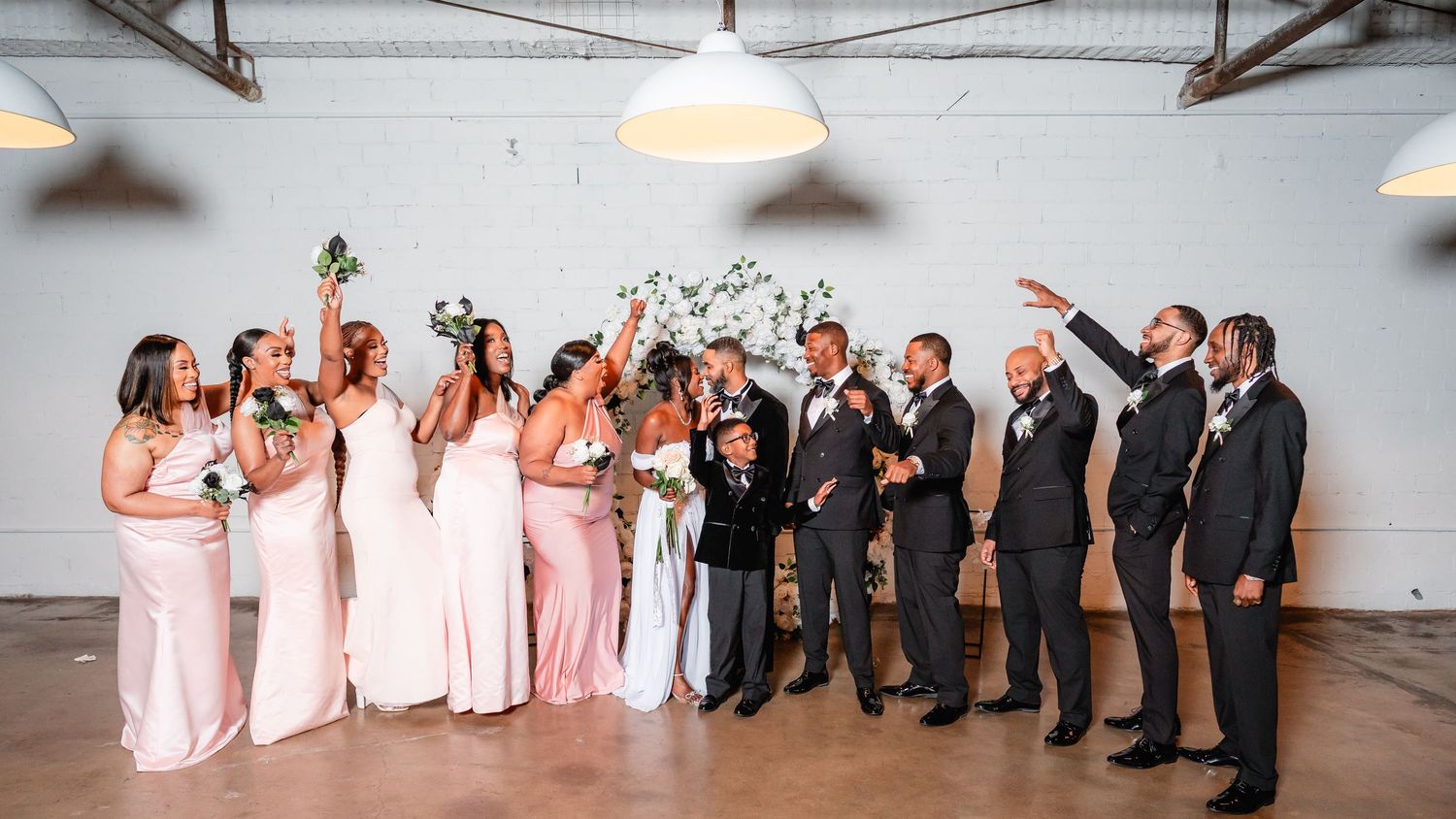 Wedding party members pose in a line inside an industrial venue with hanging lights.