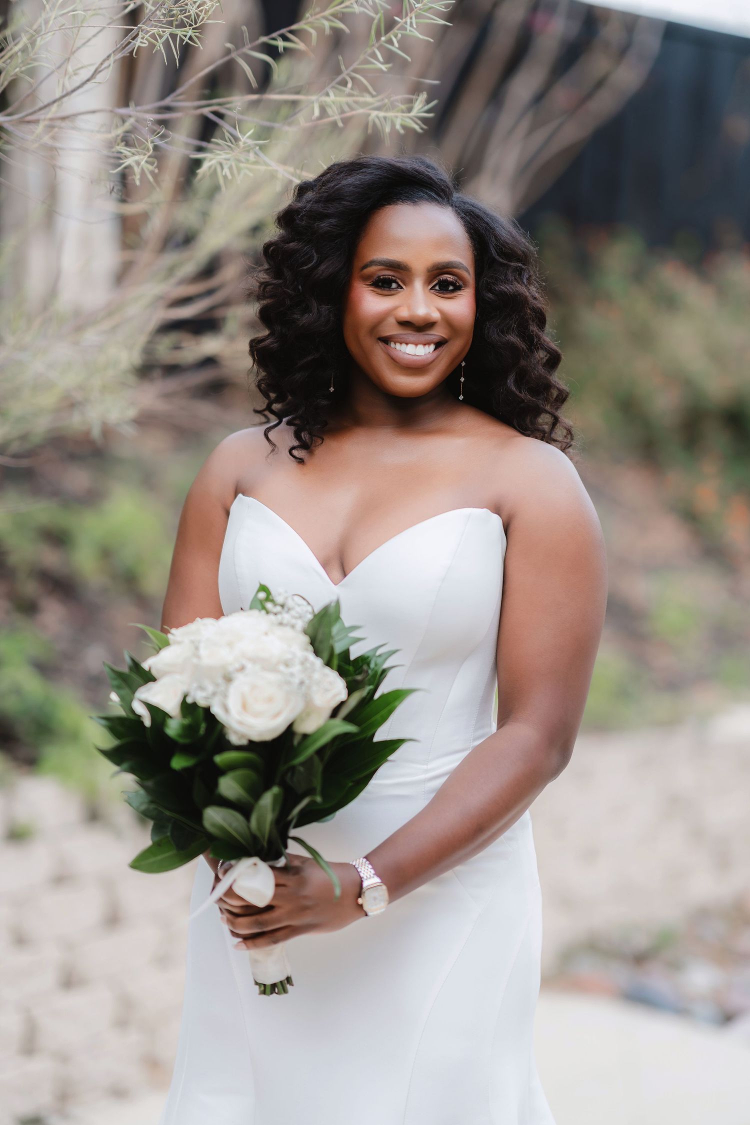 A bride in a white strapless wedding dress holds a classic white rose bouquet against a natural outdoor backdrop.