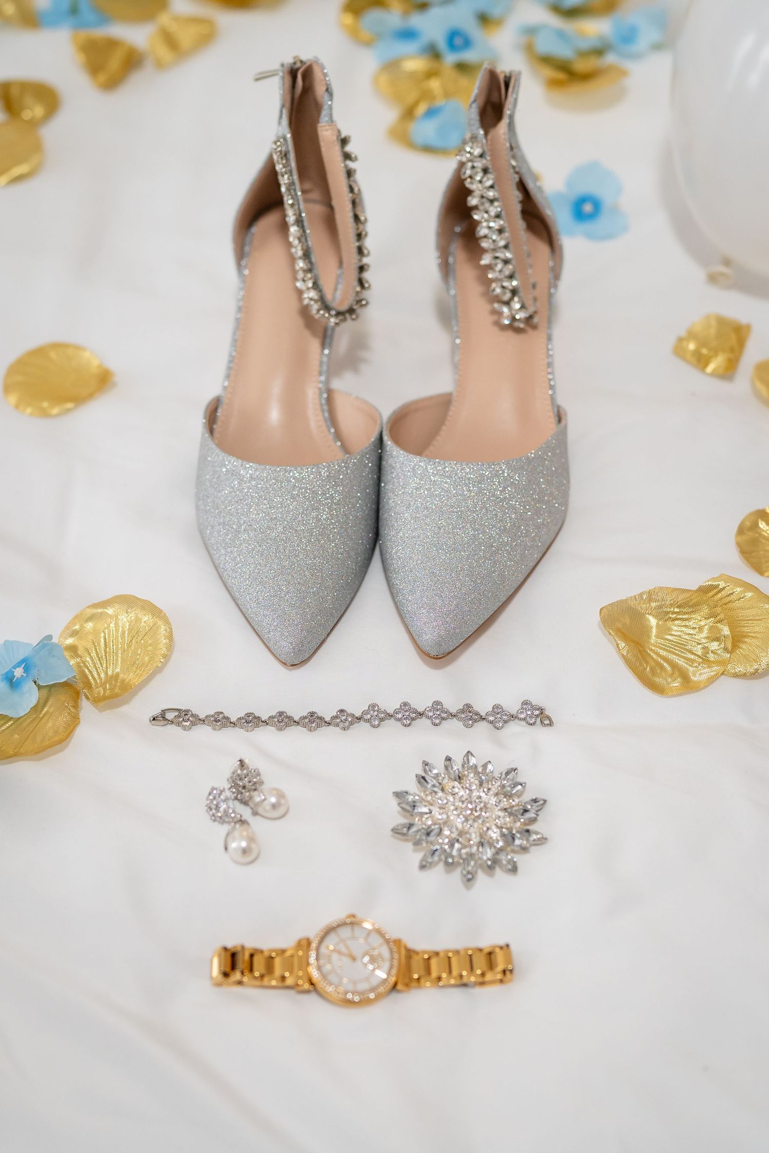 Wedding accessories including silver glitter shoes, jewelry and a gold watch arranged on white fabric with blue petals.