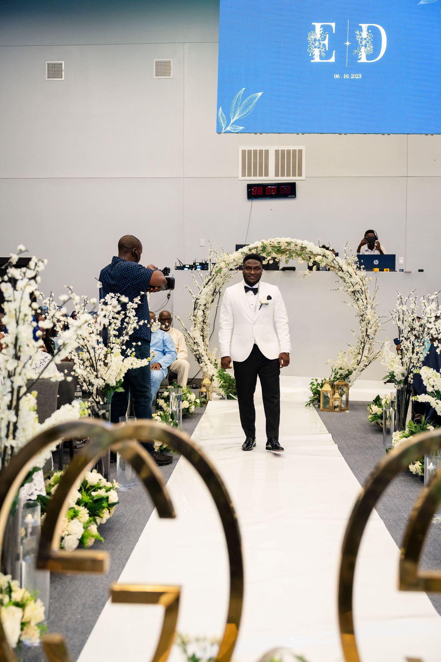 A groom stands at a floral arch decorated ceremony site with white flowers.