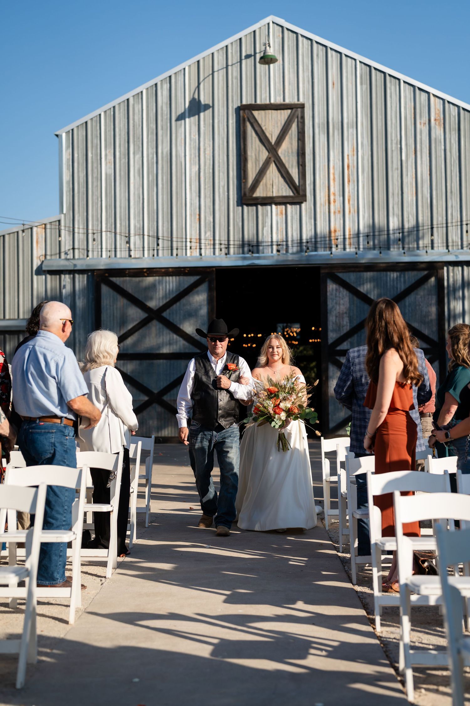 Wedding guests mingle at an outdoor venue with white chairs arranged in front of modern barn-style buildings.
