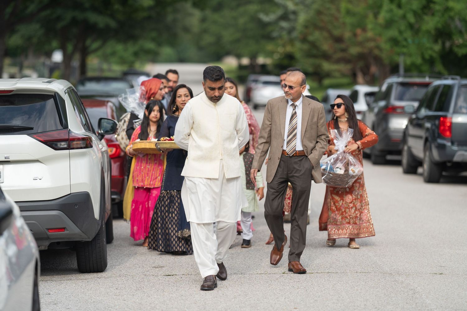 Wedding guests walking outside on a paved path surrounded by parked cars.