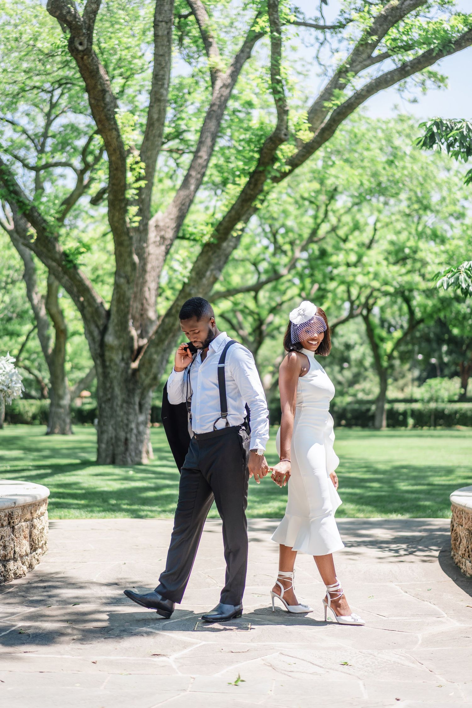 A couple poses playfully on a garden path under a canopy of green trees.