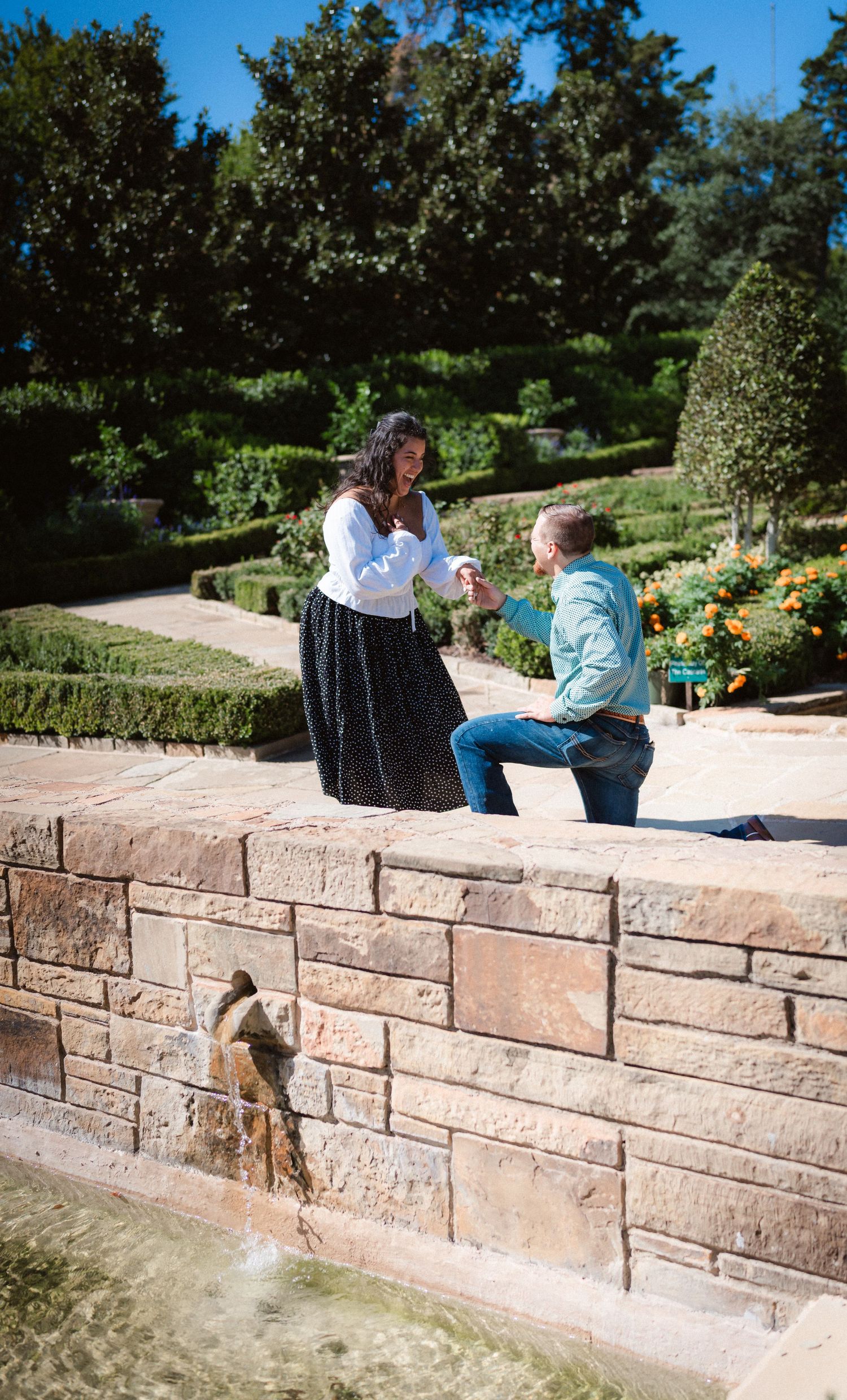 A proposal moment captured in a formal garden with stone walls and fountains on a sunny afternoon.