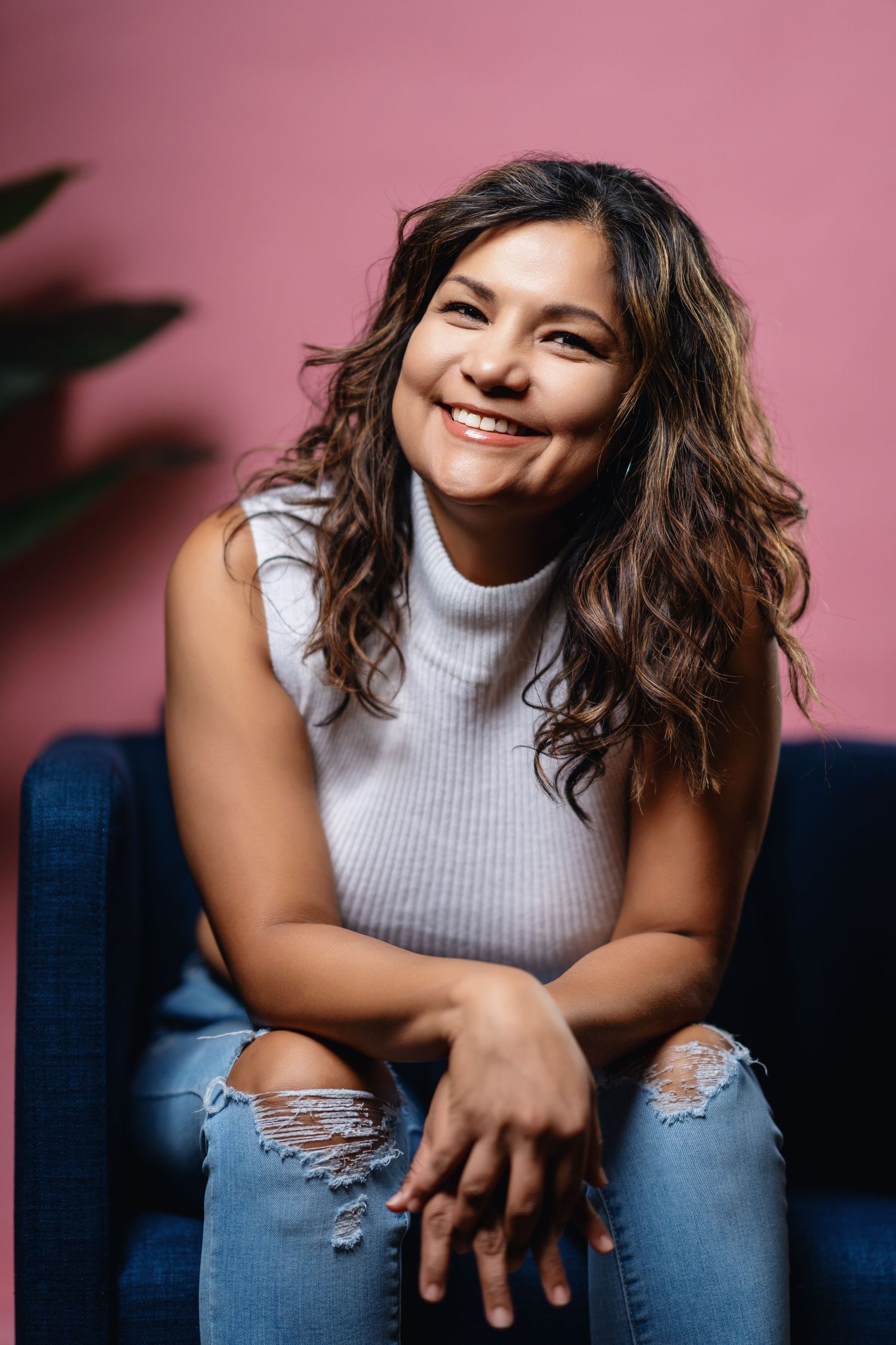Person in ripped jeans and sleeveless turtleneck poses against pink backdrop with natural curly hair and casual style.