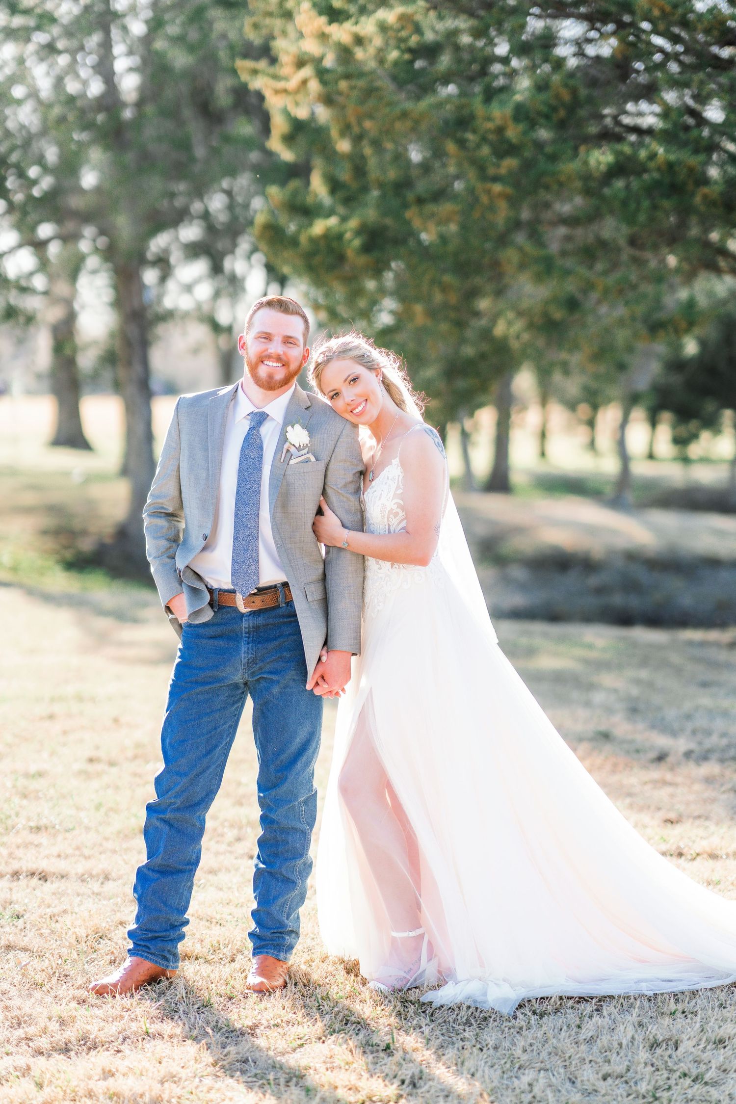A couple poses in a sunlit field, with the bride in white gown and groom in casual suit and jeans.