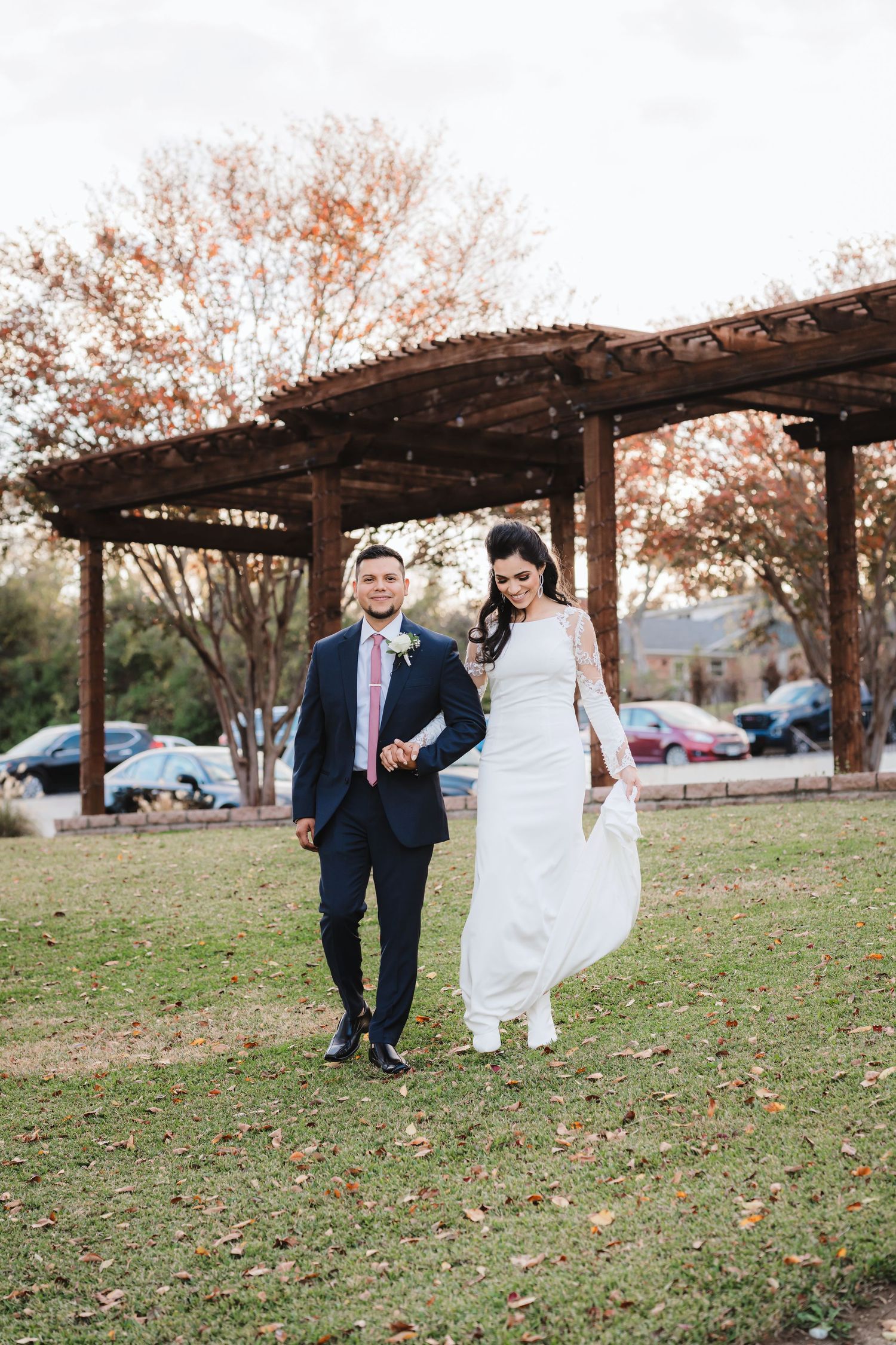 Newlyweds walk playfully under wooden pergola in outdoor garden setting.