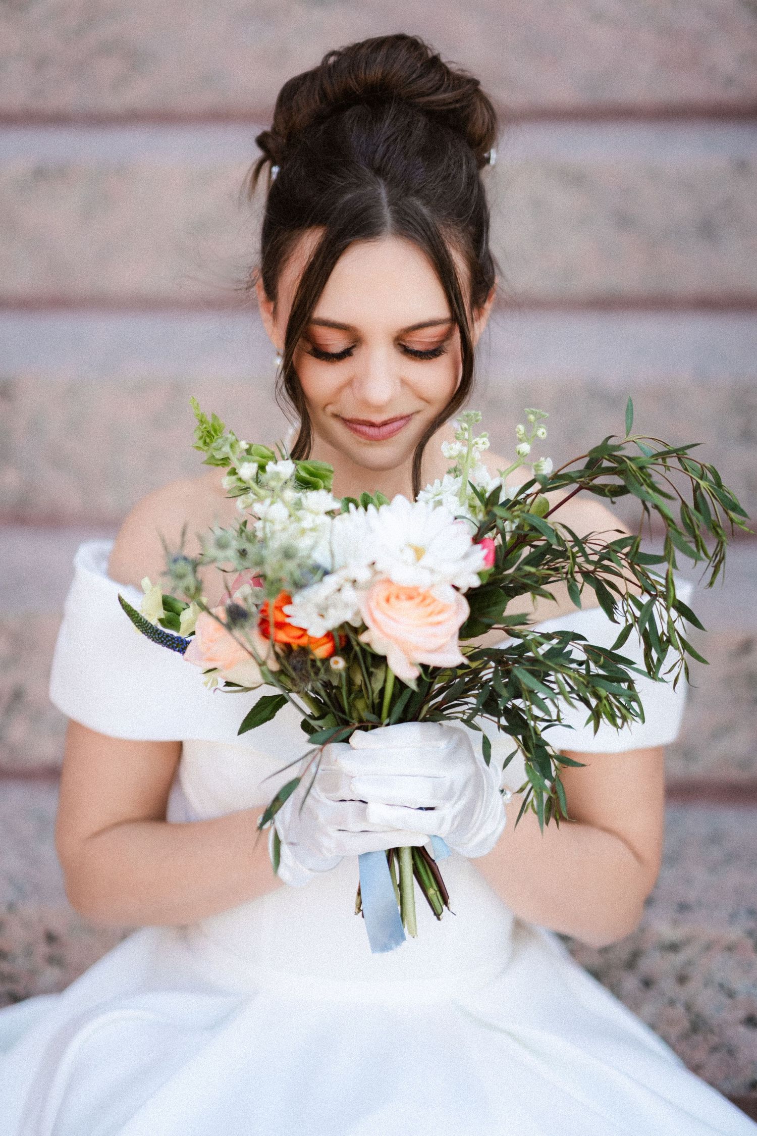 A bride in a white dress smiles while holding a delicate bouquet of peach and white flowers with greenery.
