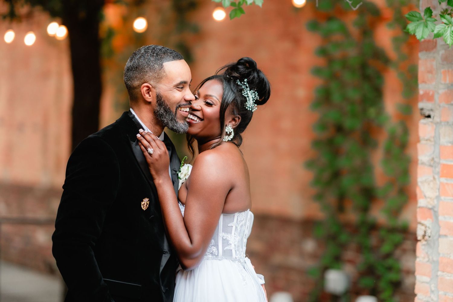 Romantic wedding couple embraces in a rustic brick courtyard with twinkling lights and ivy in the background.