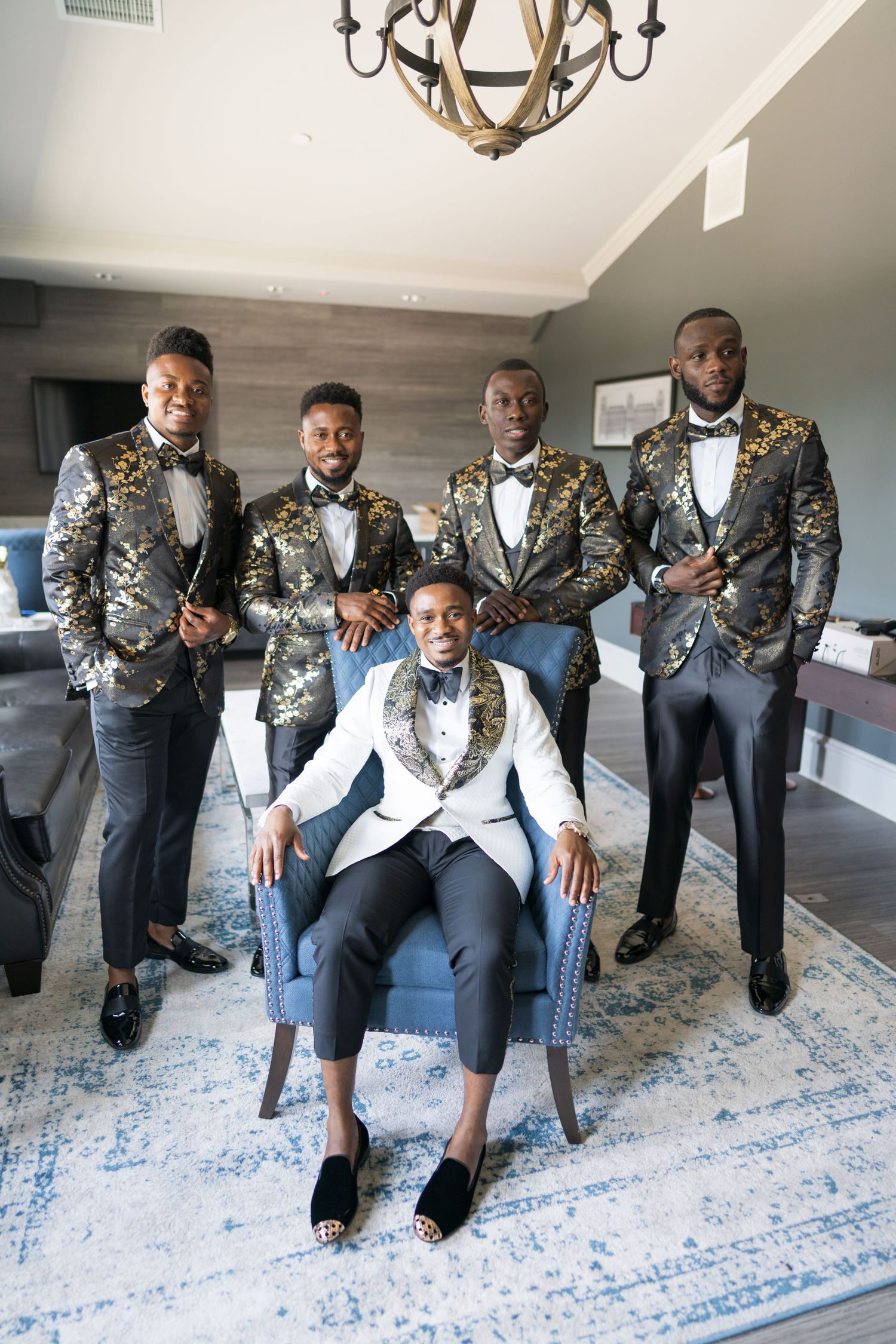 A wedding party poses in matching gold brocade tuxedo jackets and bow ties with one member seated in a gray chair.