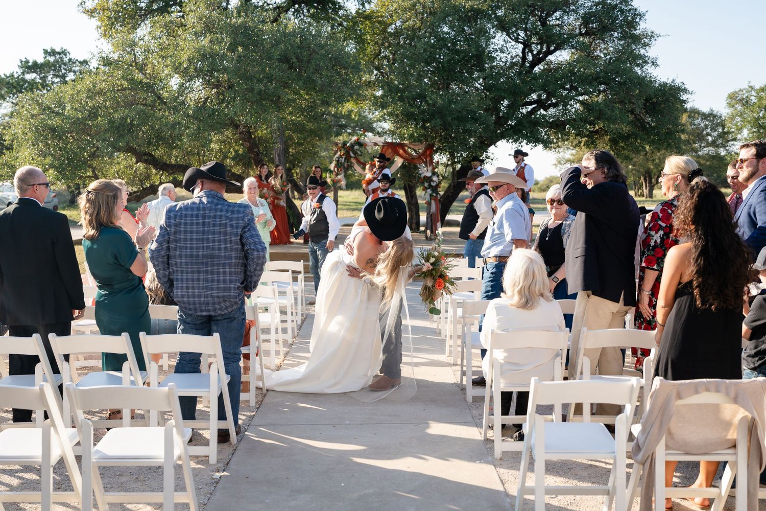White chairs set up outdoors for a wedding ceremony under large shade trees.
