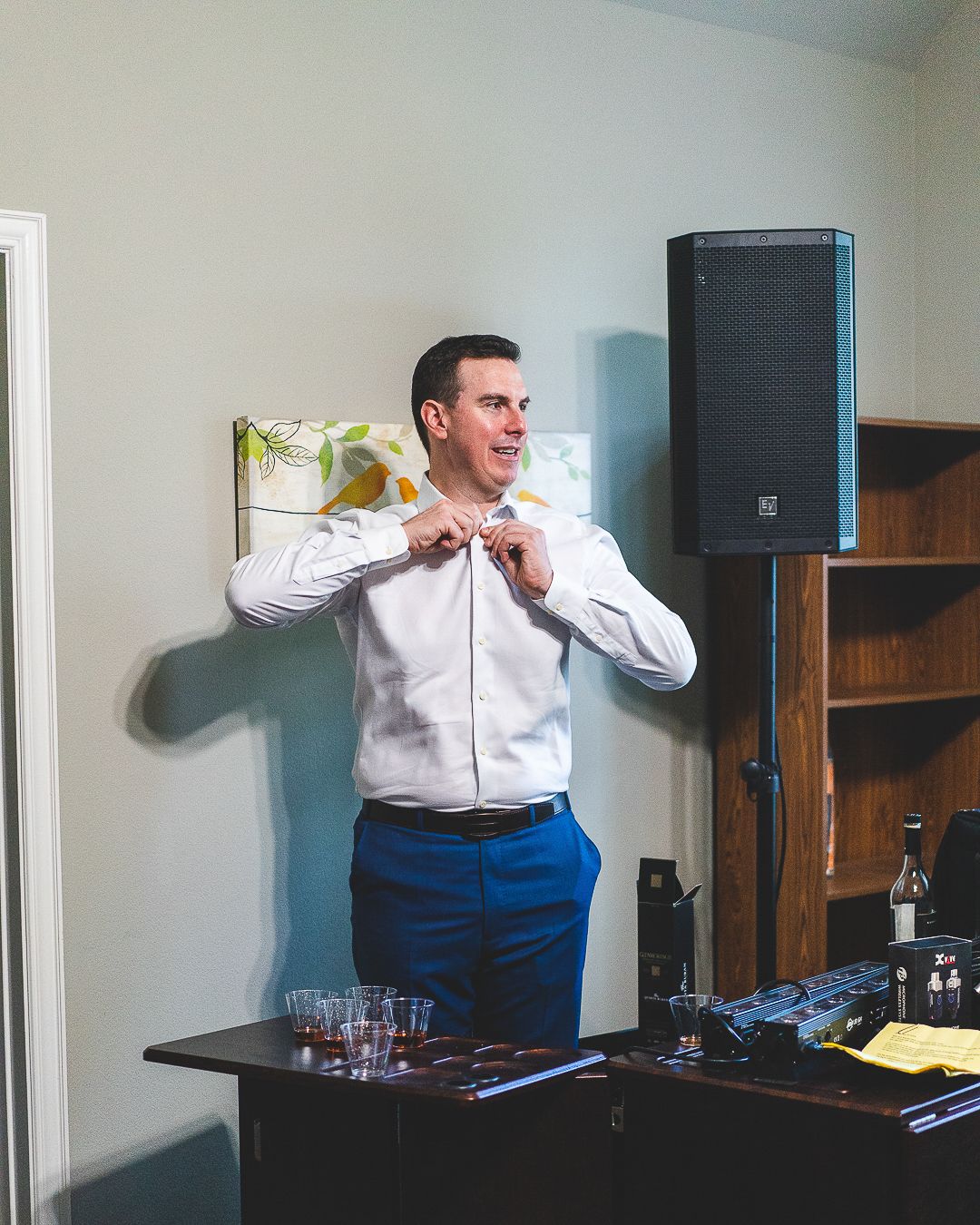 A person in a white dress shirt and blue pants adjusts their tie while standing near audio equipment.