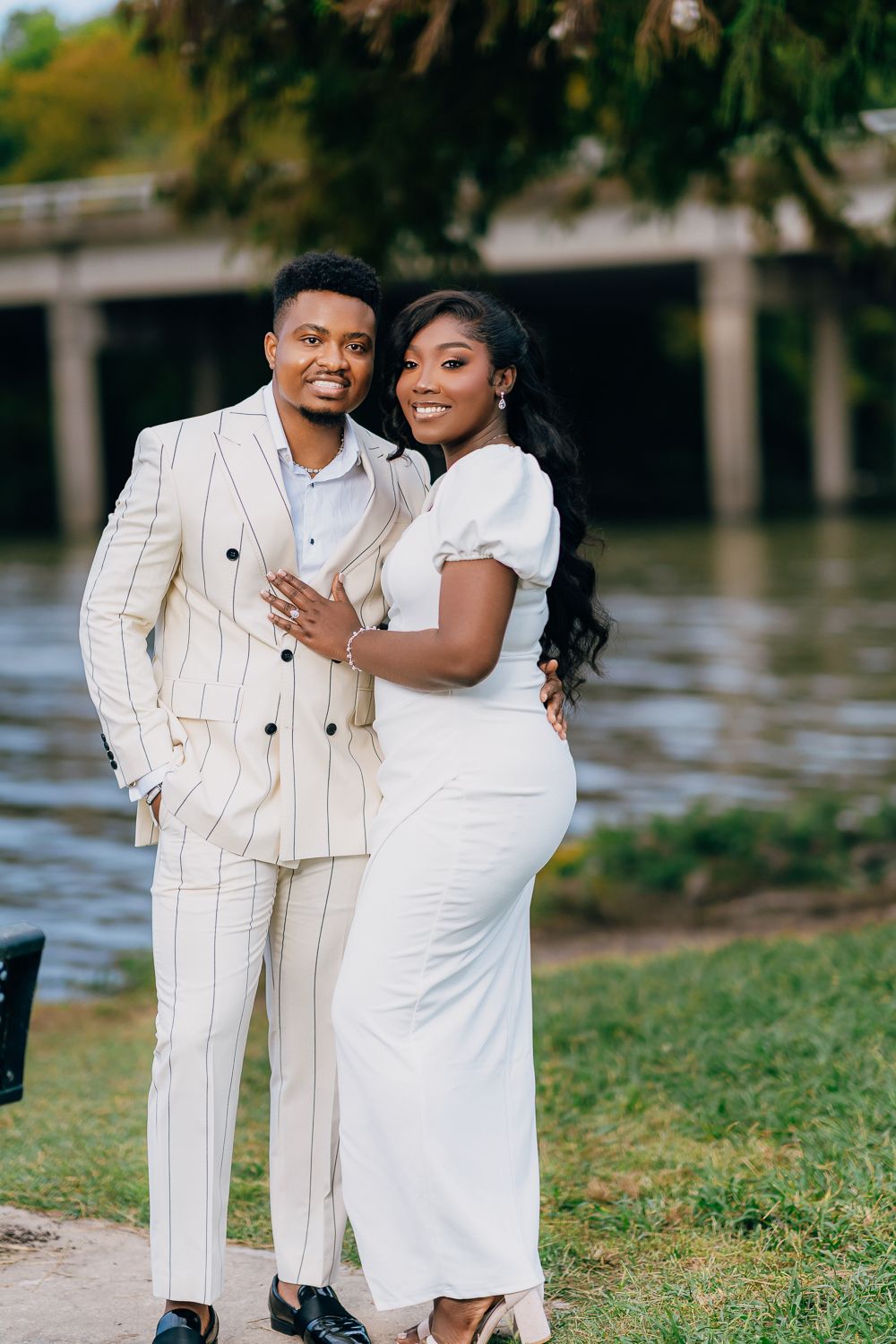 A couple in matching white outfits poses together by a waterfront during a formal outdoor photoshoot.