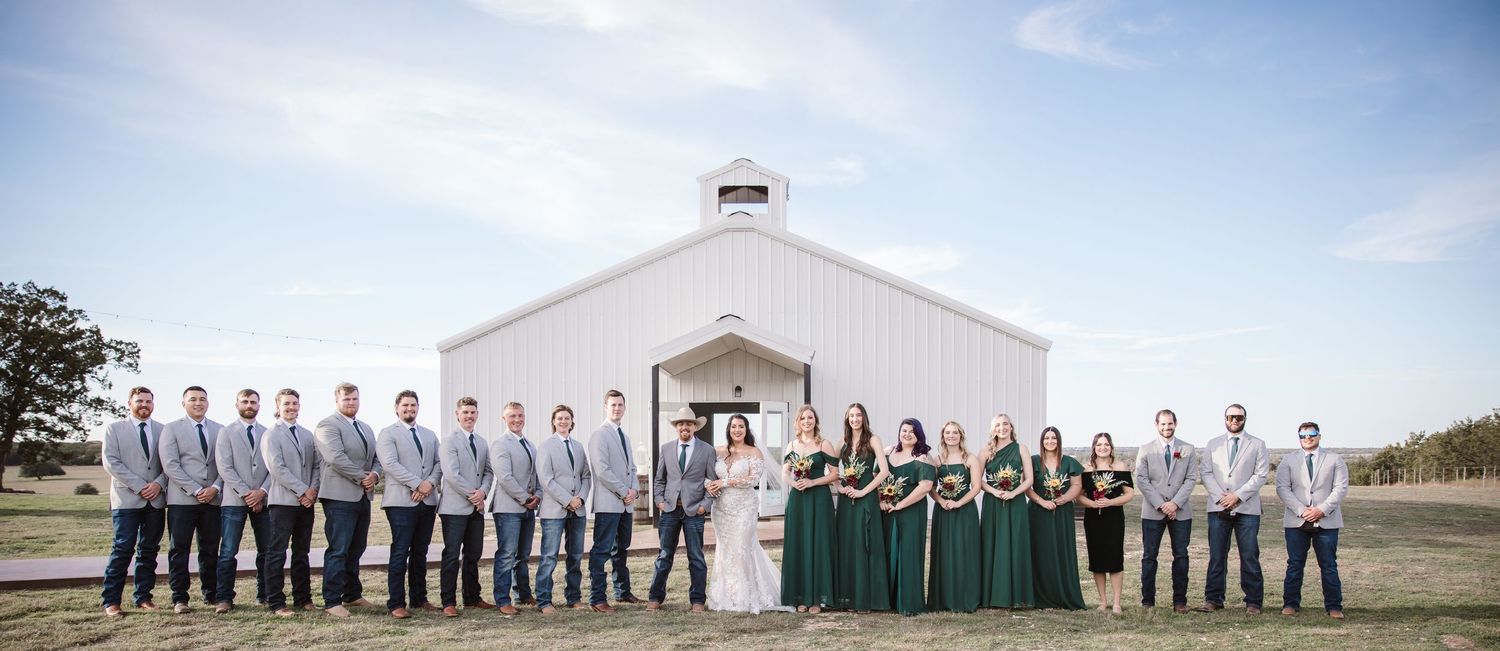 Panoramic view of a wedding party lined up outside a white barn-style building under blue skies.