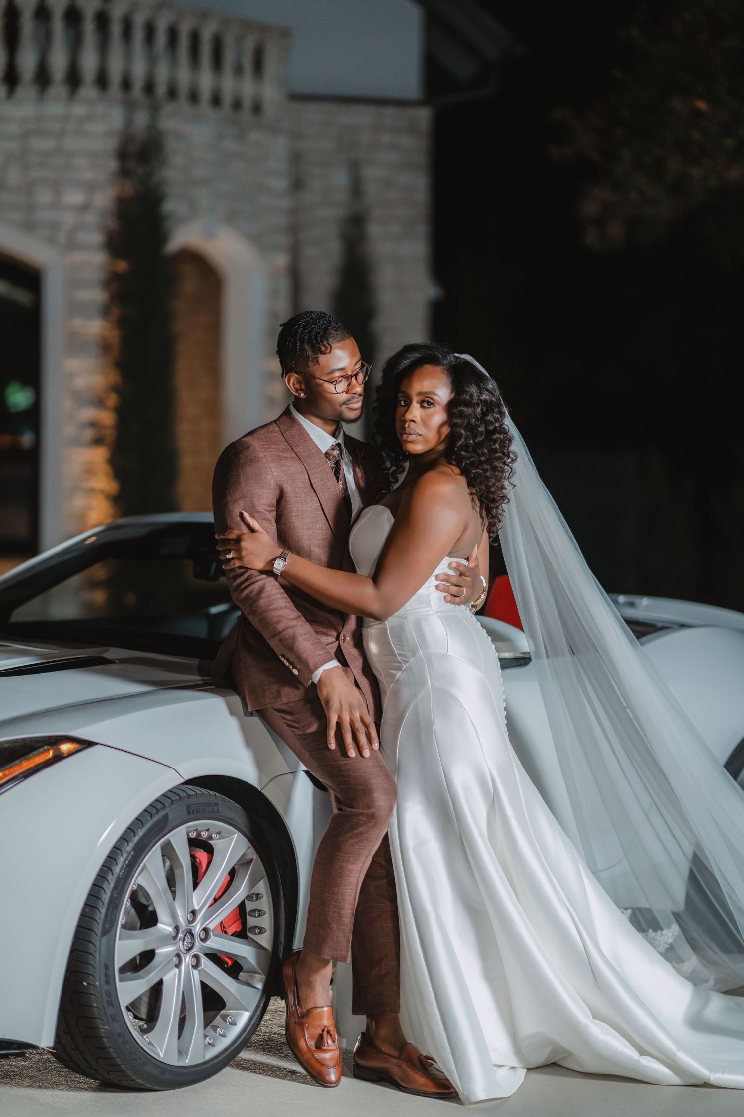 Elegant wedding couple poses by a luxury car in formal attire.