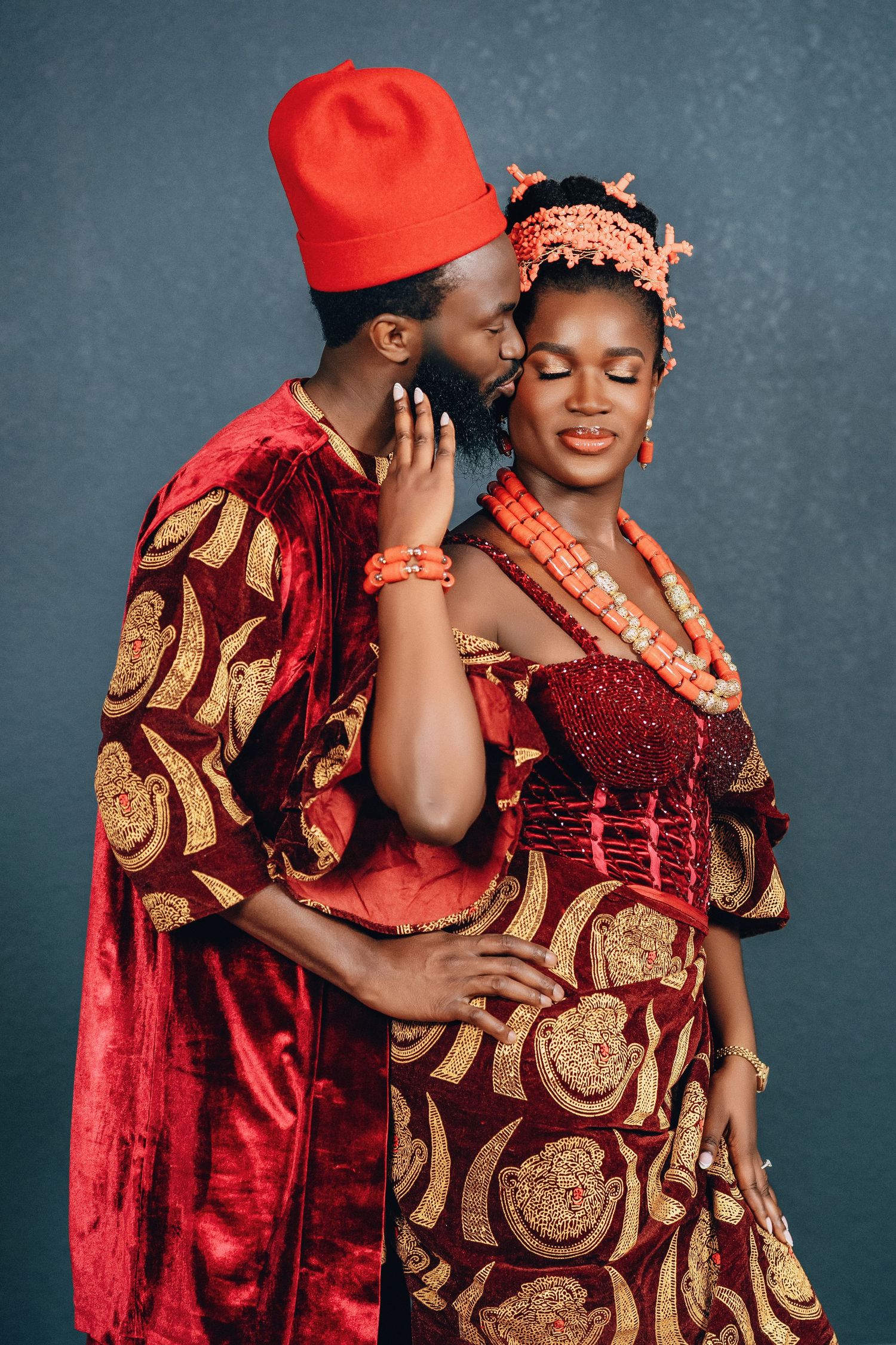 Romantic couple in traditional Nigerian wedding attire embracing against blue backdrop.