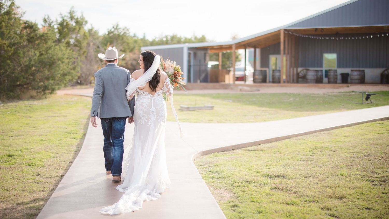 Wedding party members walk together across a grassy field outside a rustic venue.
