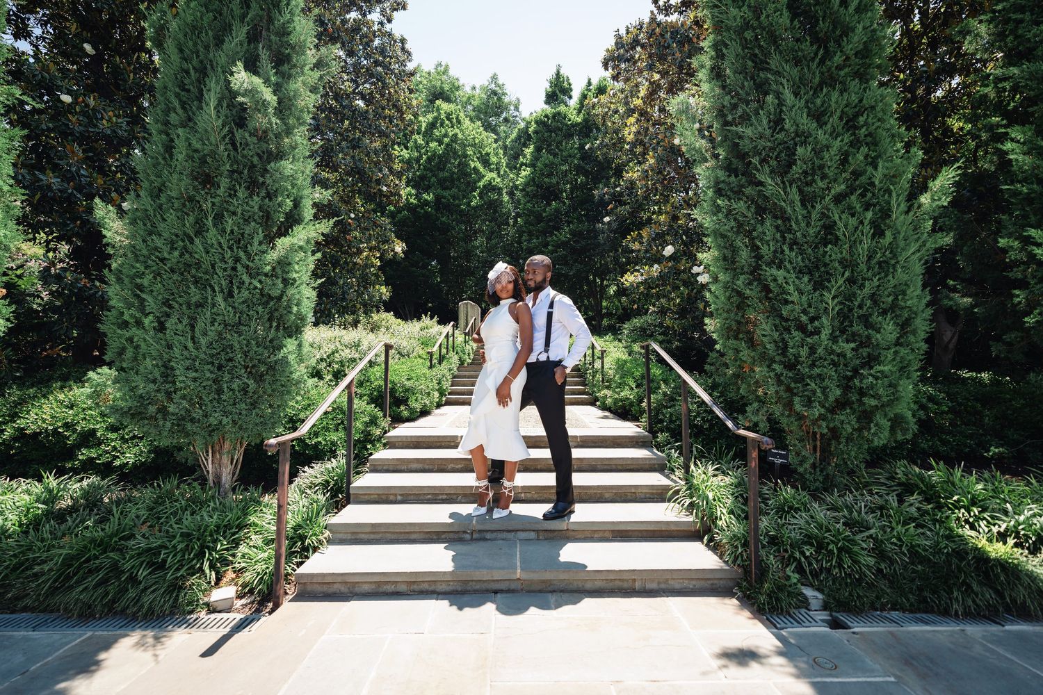 A couple sits together on stone steps surrounded by tall evergreen trees in a formal garden.
