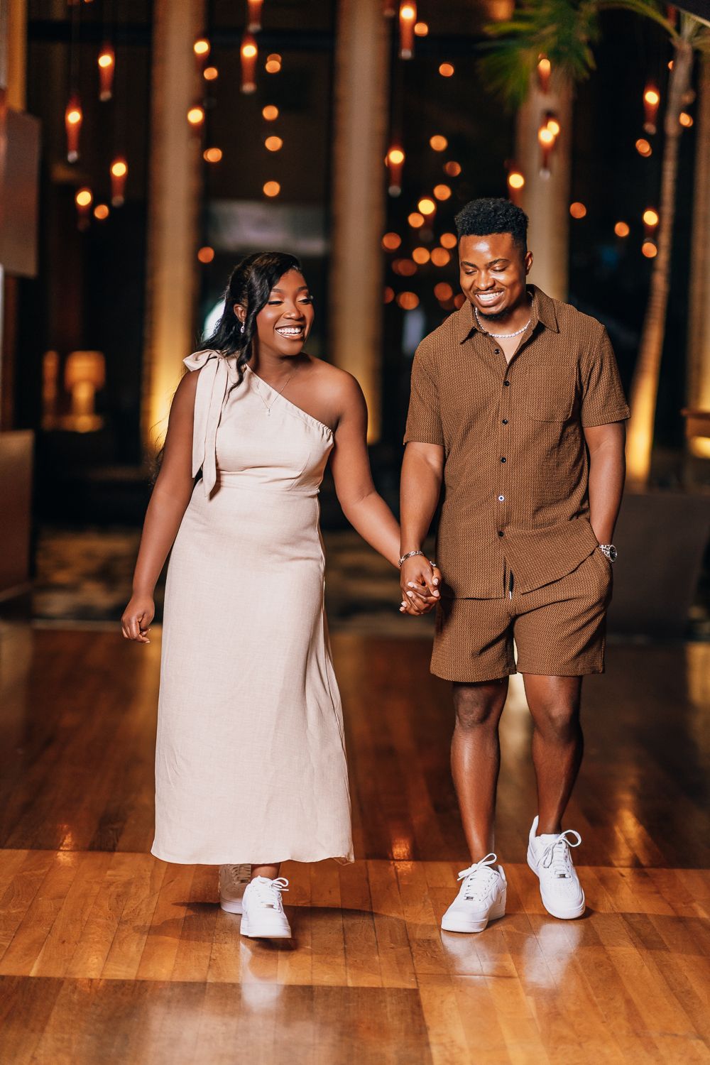 A couple walks hand in hand on a wooden floor under romantic string lights during their engagement session.
