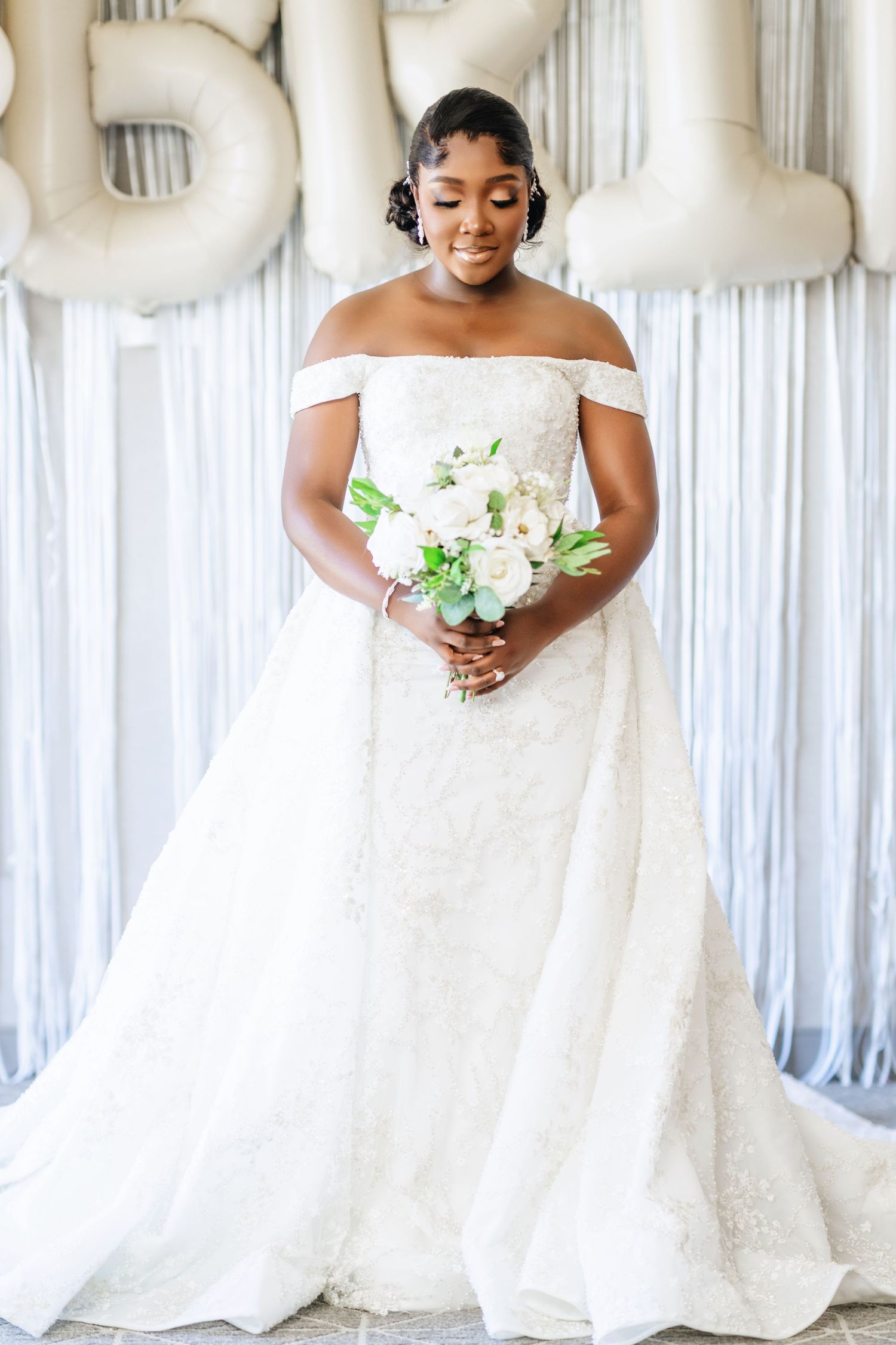 A bride in an elegant white off-shoulder wedding gown holds a delicate white bouquet against a light backdrop.