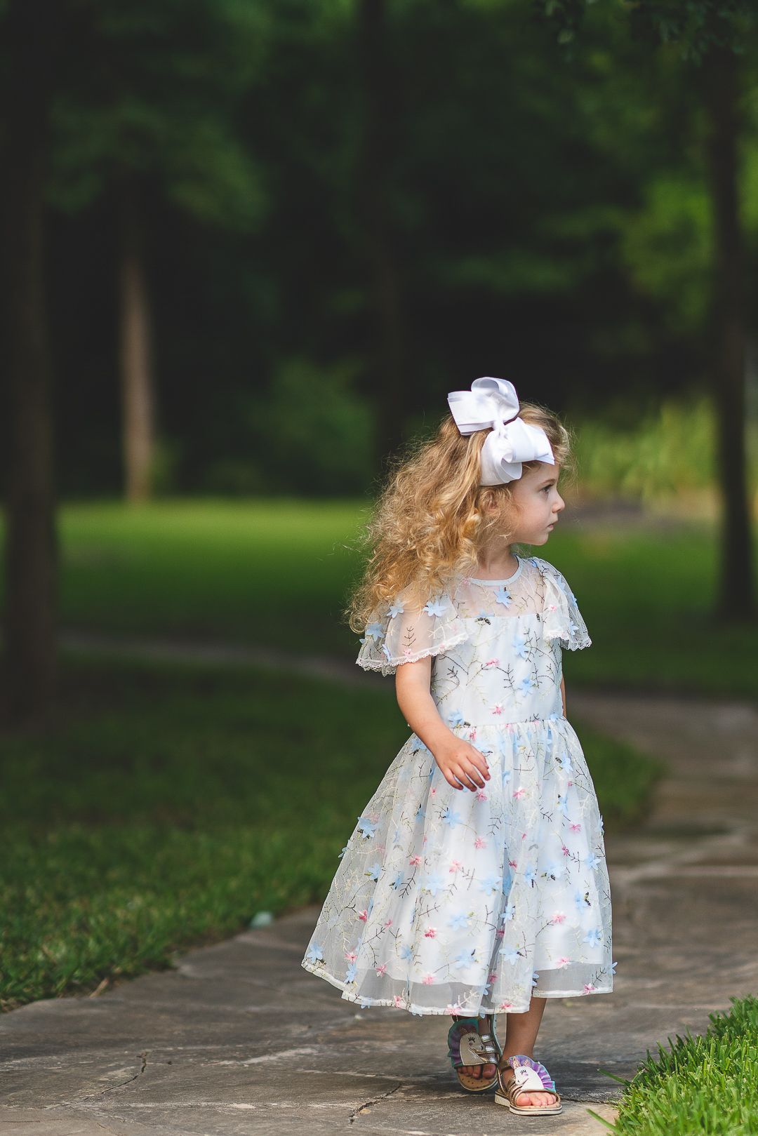 A young flower girl in a floral dress walks along a garden path.