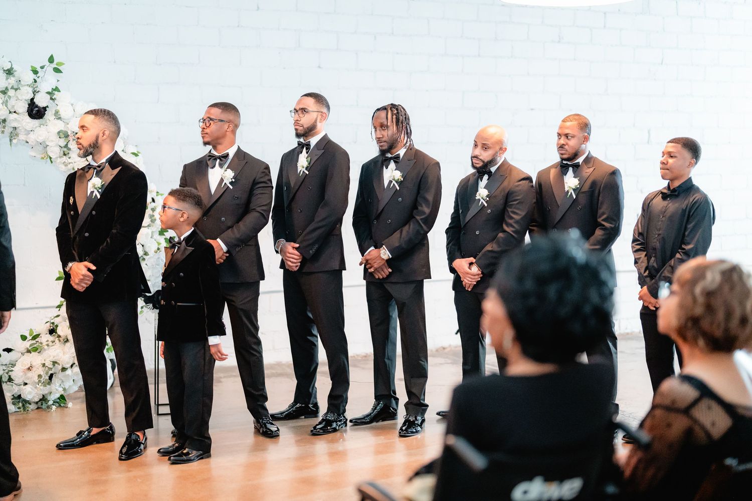 Groomsmen in dark suits stand in a line at a wedding ceremony.