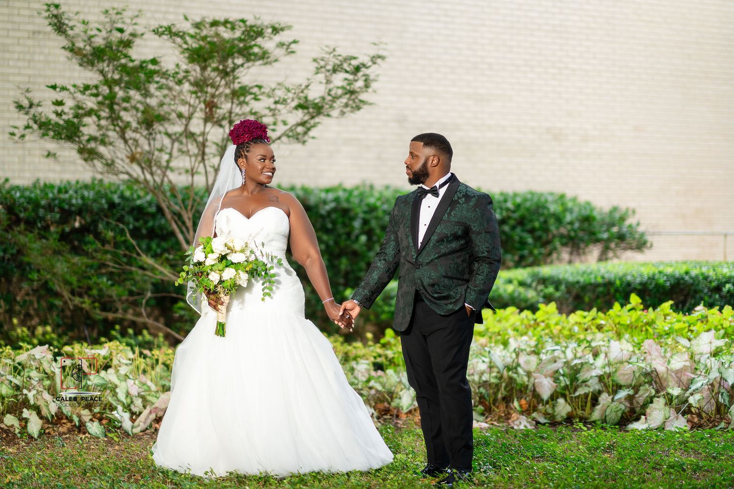A couple in formal wedding attire poses together in a lush garden setting with white flowers and greenery.