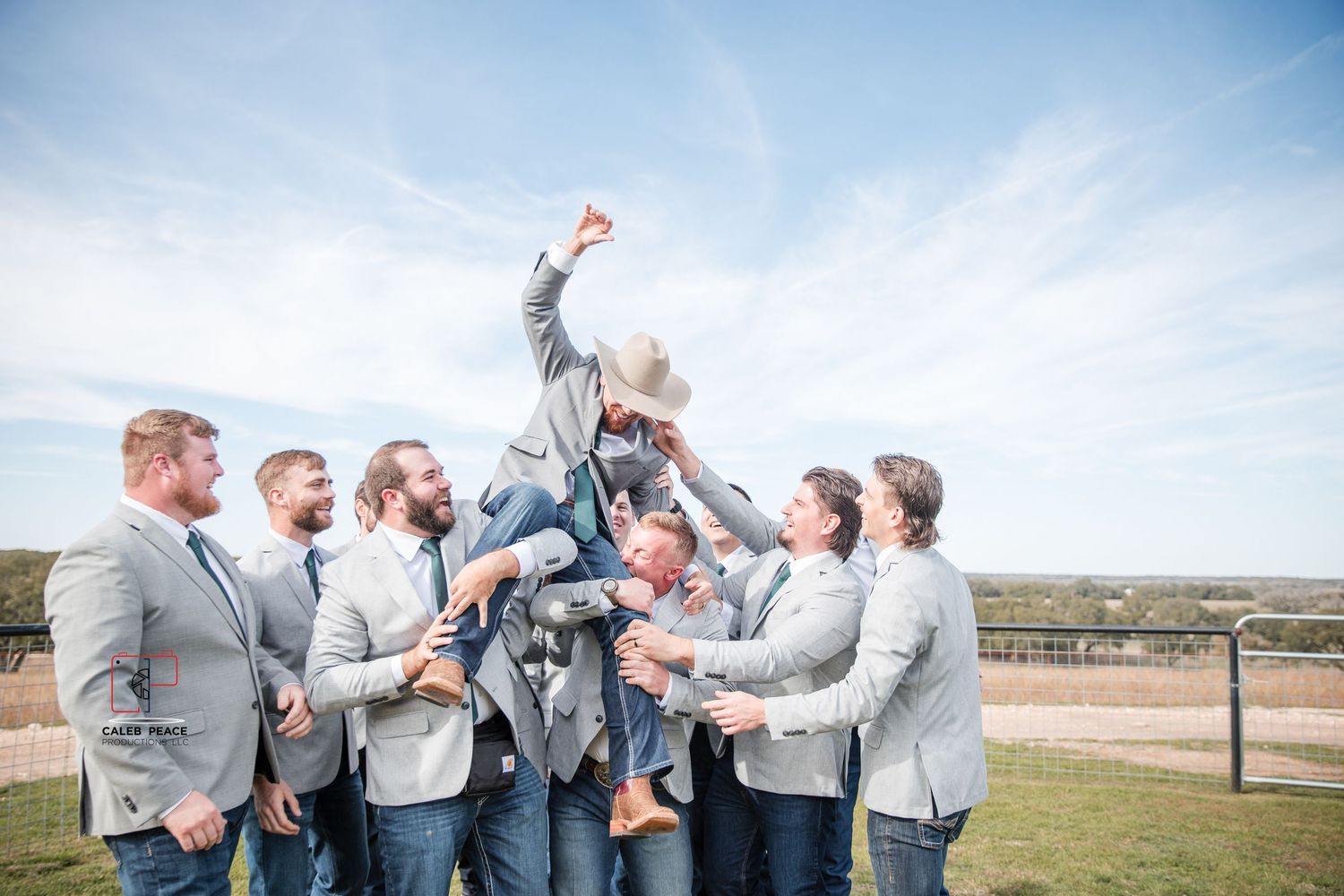 Groomsmen in gray suits celebrate joyfully outdoors against a bright blue sky.