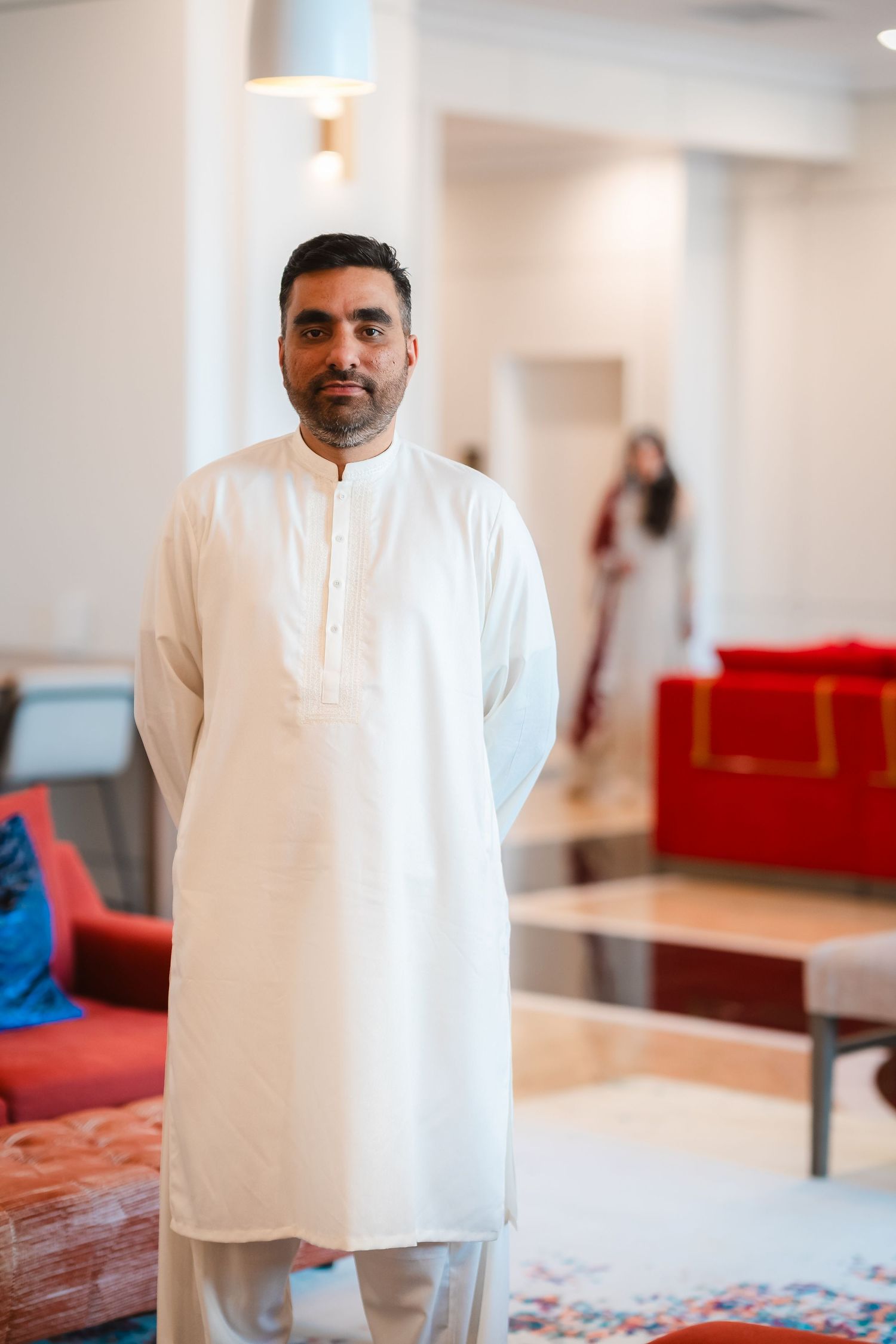 A person wearing traditional white cultural attire stands in an indoor space with red seating.