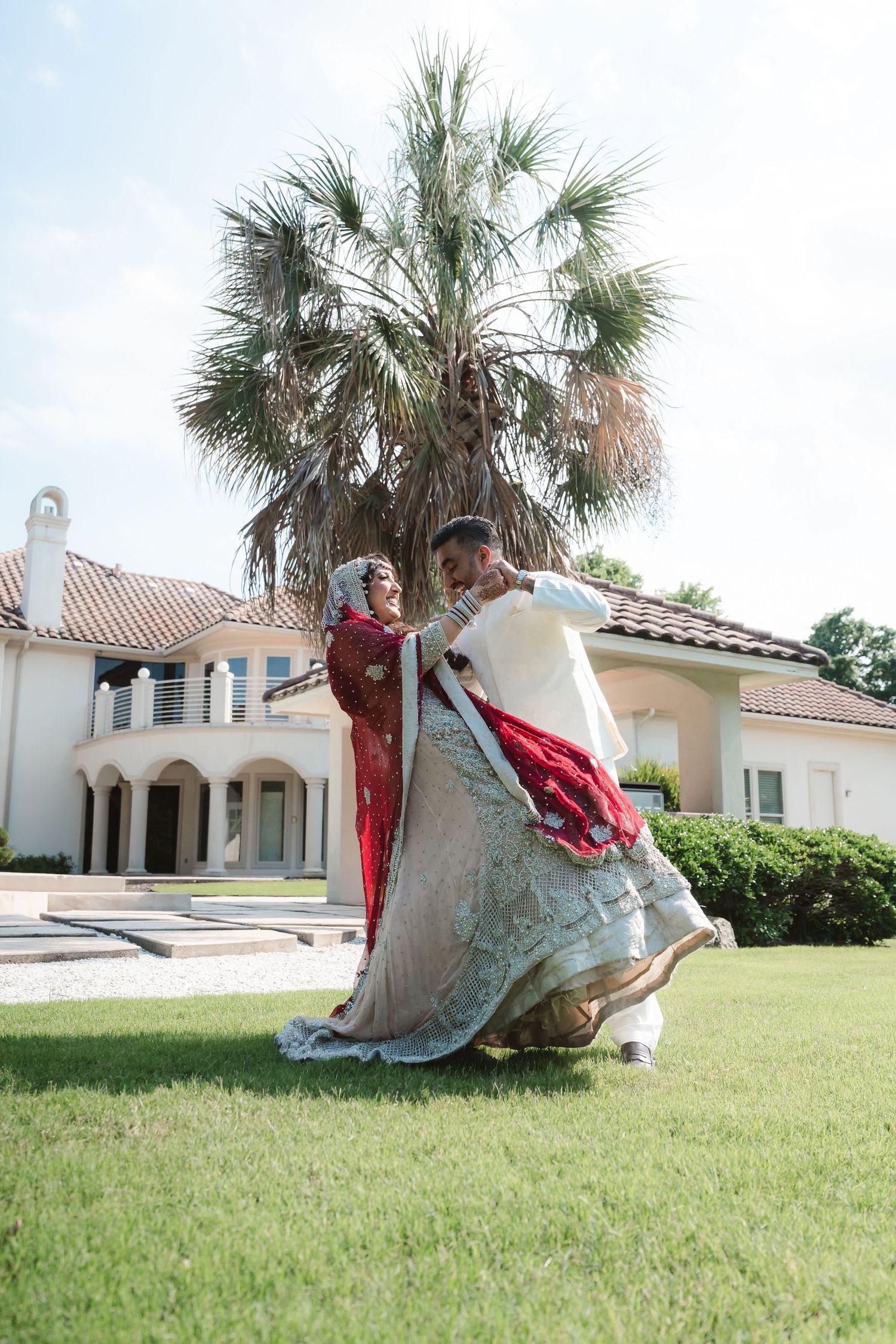 Wedding couple dancing under palm trees in front of Mediterranean-style mansion.