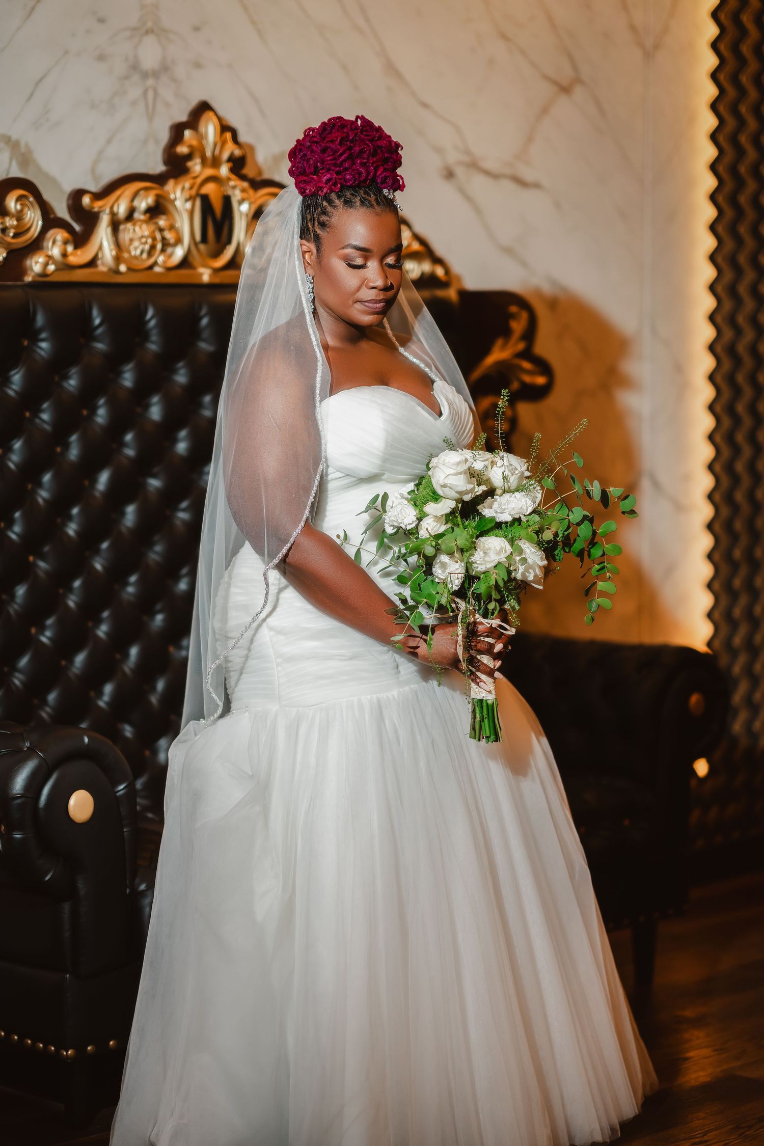 Bride in elegant white ballgown holds bouquet while posing against luxurious tufted backdrop.