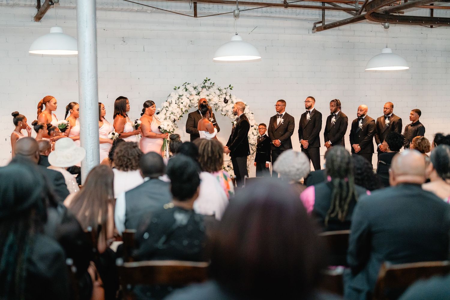 Wedding ceremony in modern industrial space with floral arch and gathered wedding party.