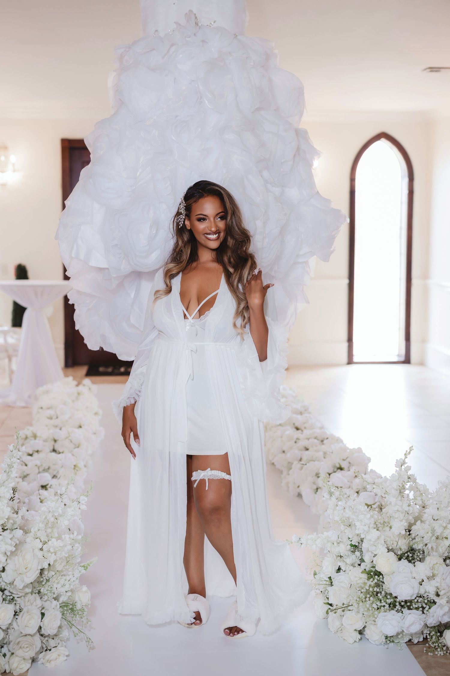 Bride in short white dress and robe poses with dramatic floral backdrop and white decor.