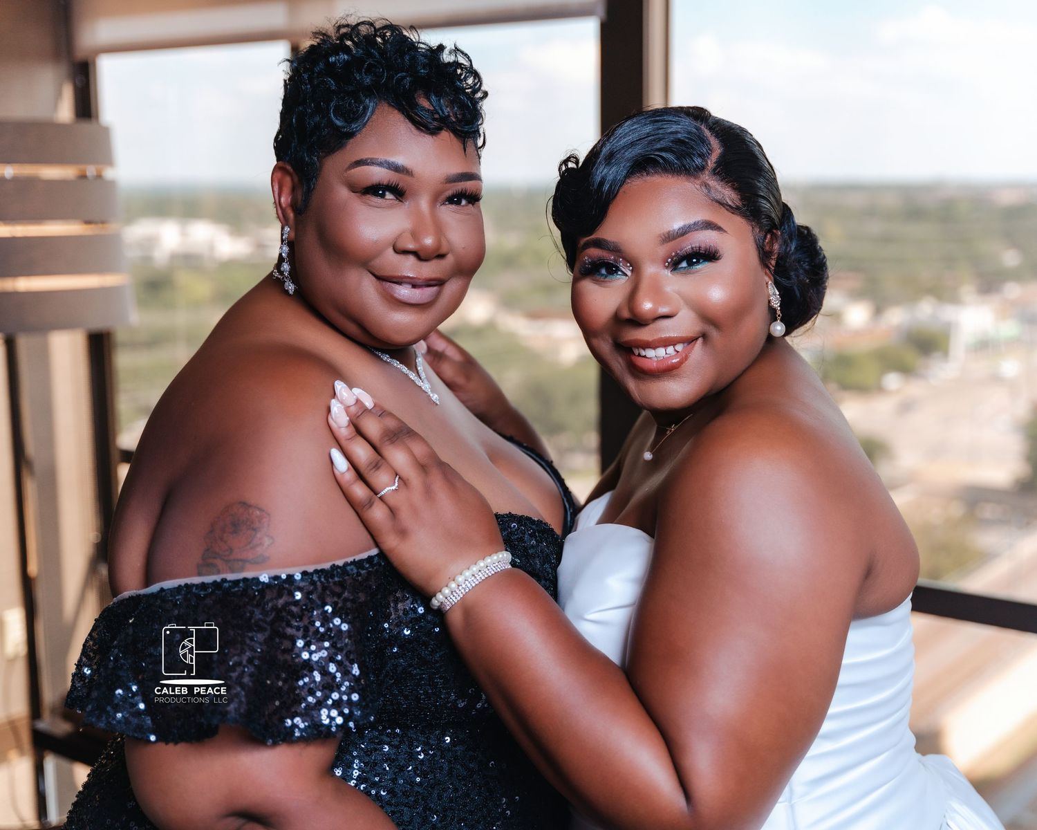 Wedding party members in sparkling black gowns and white dress pose together by large windows.