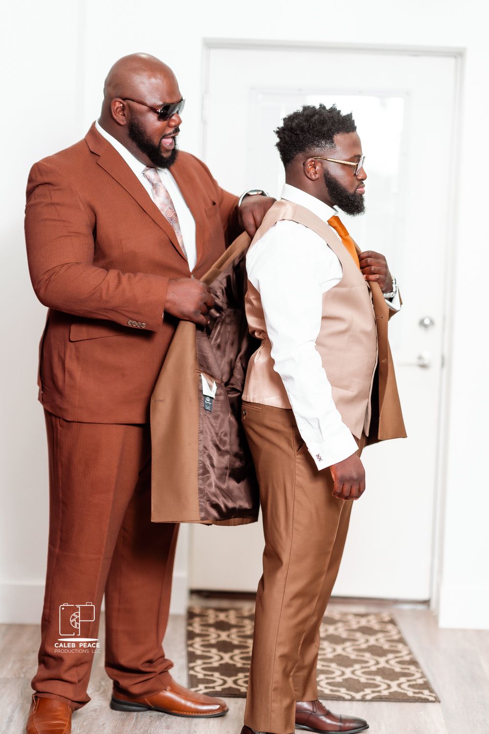 Two stylishly dressed people in brown and tan suits help with fitting adjustments in a bright room.