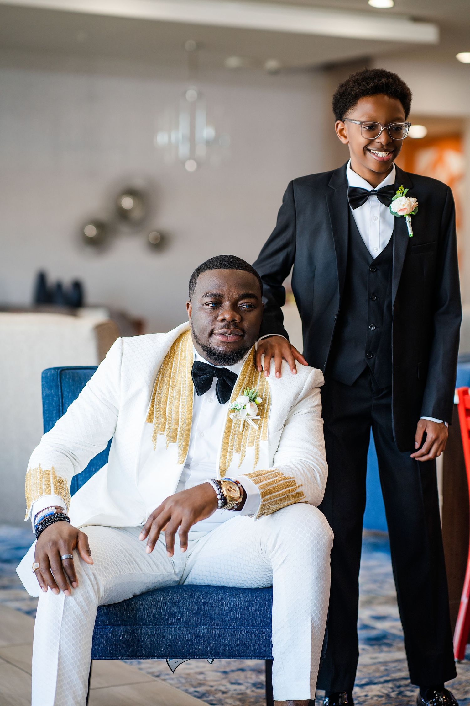 Wedding party member in crisp white suit poses on blue couch for formal portraits.