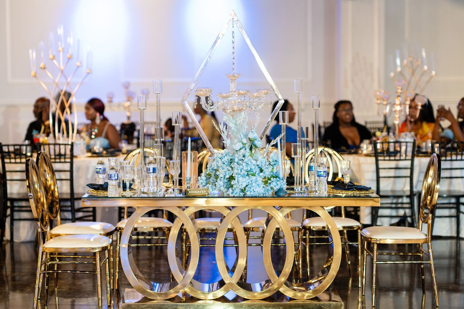 An elegant wedding reception venue features crystal chandeliers and gold chairs with blue accent lighting.