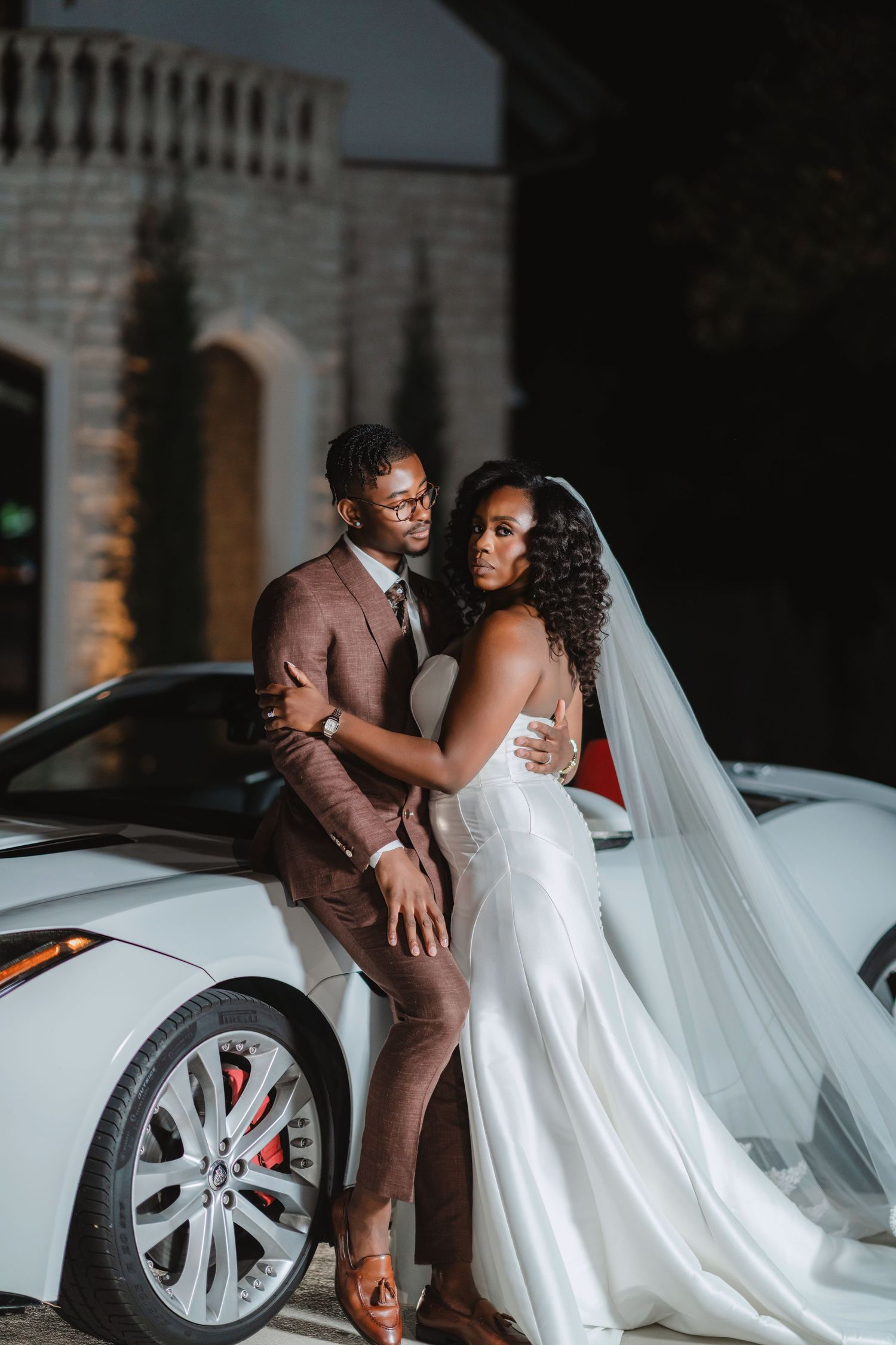 Romantic wedding photoshoot featuring a couple posing by a white luxury vehicle at dusk.