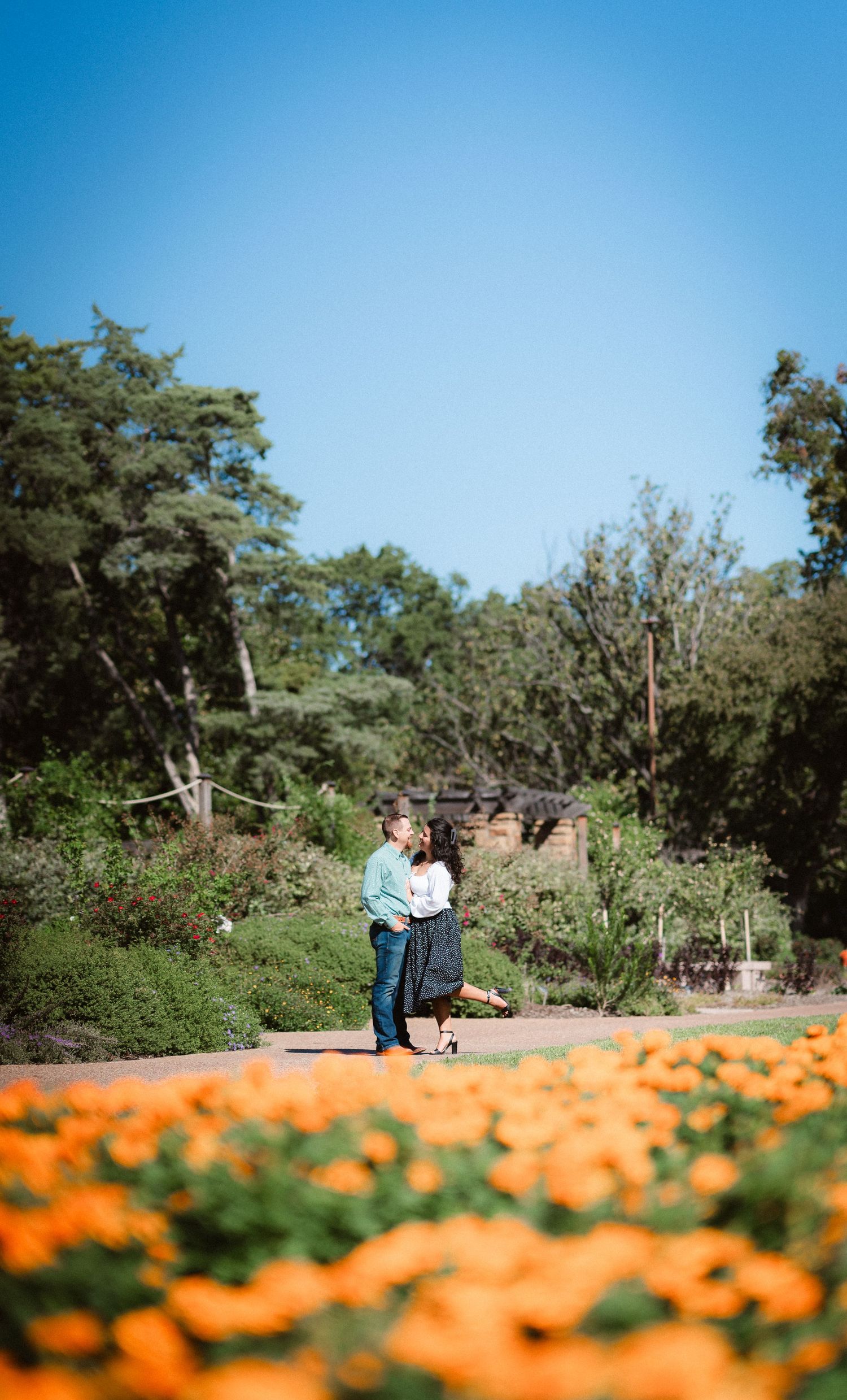 A couple stands together in a garden with vibrant orange flowers blooming in the foreground.