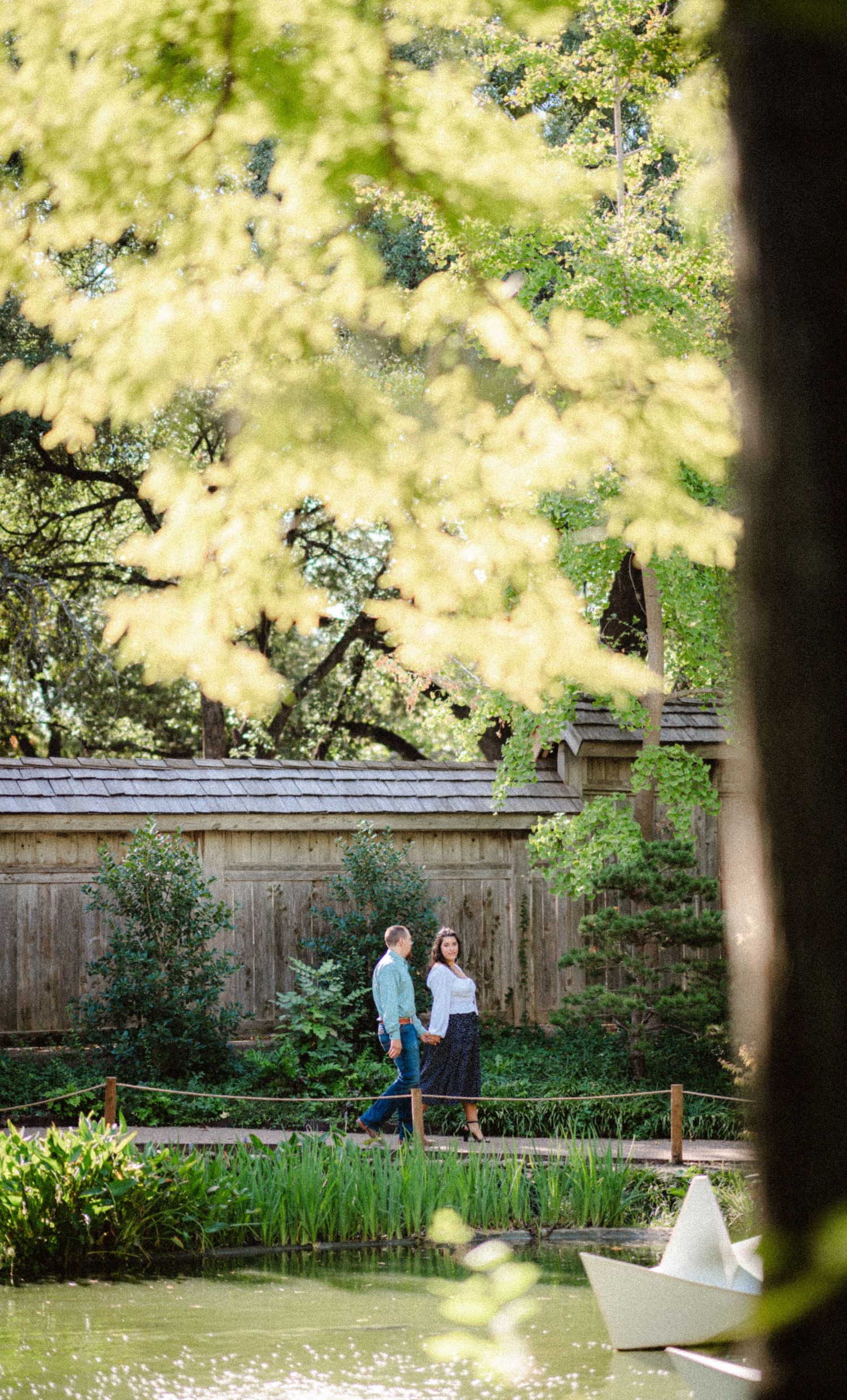 A couple stands near a pond with a paper boat in a tranquil garden setting framed by golden foliage.