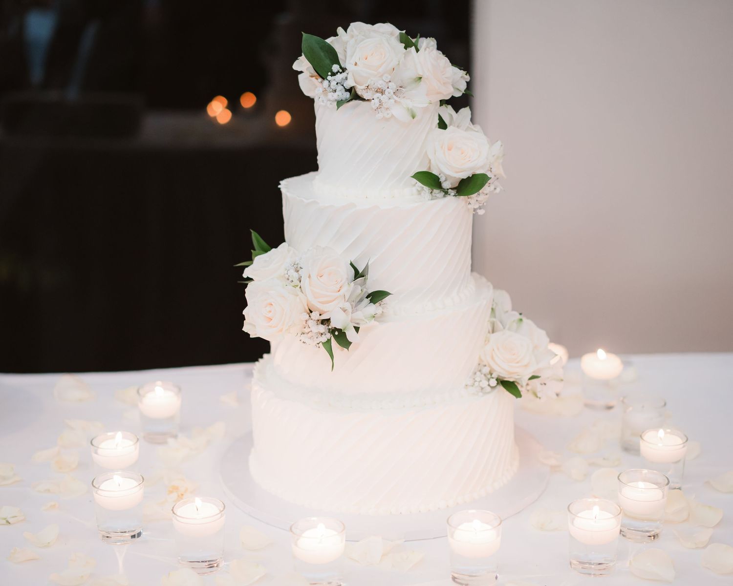 Elegant white wedding cake decorated with delicate flowers and surrounded by glowing candles on a white tablecloth.