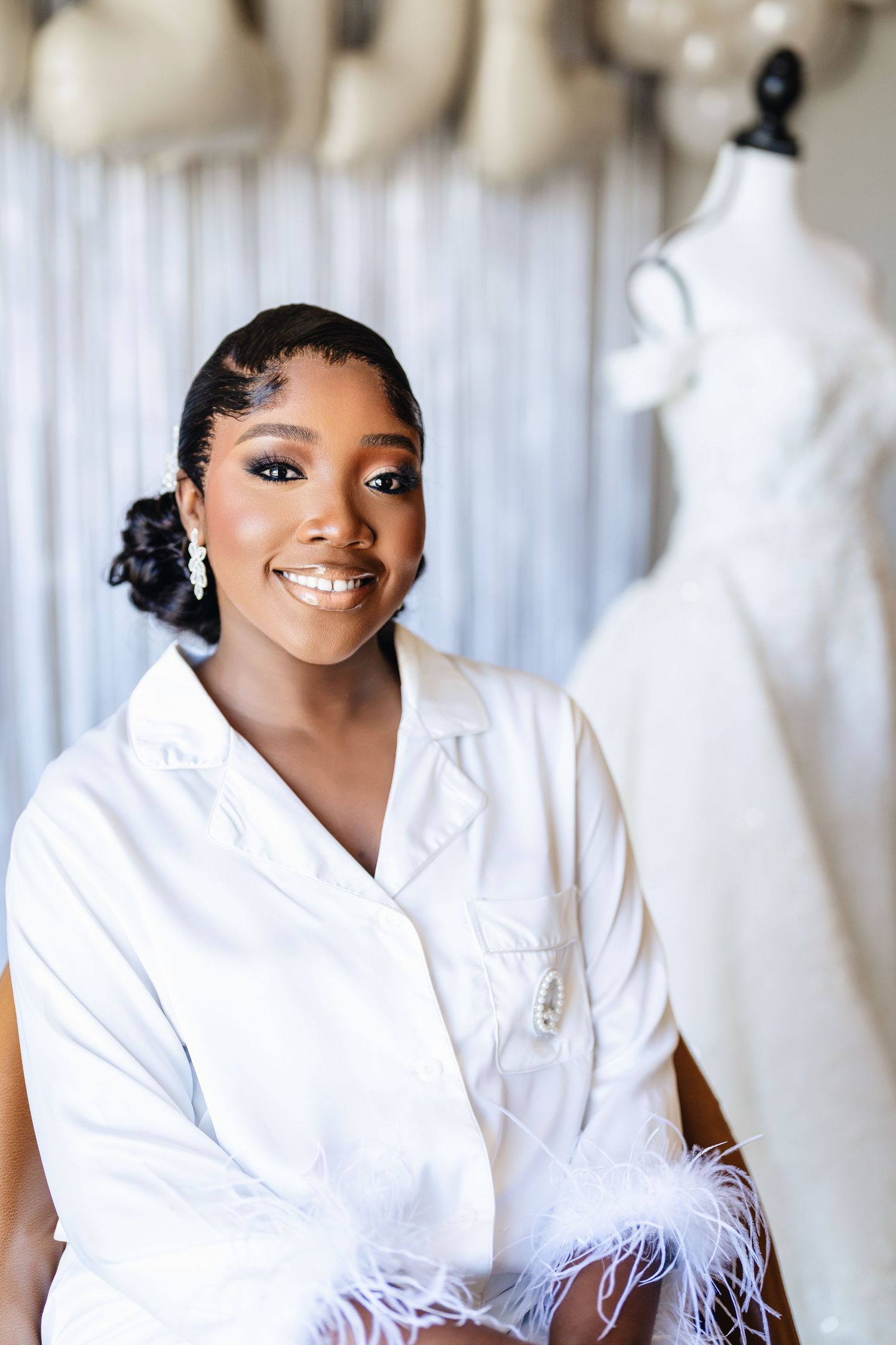 A woman wearing a white feather-trimmed robe smiles in a bridal boutique setting.