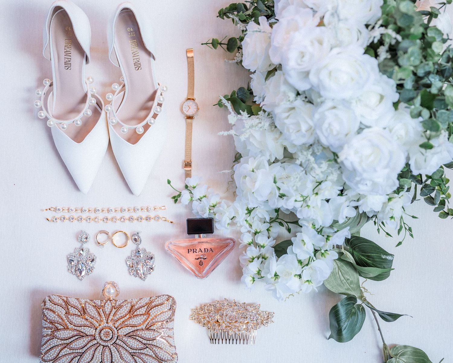 Wedding accessories flatlay with pink heels, perfume, jewelry and white flowers on light background.