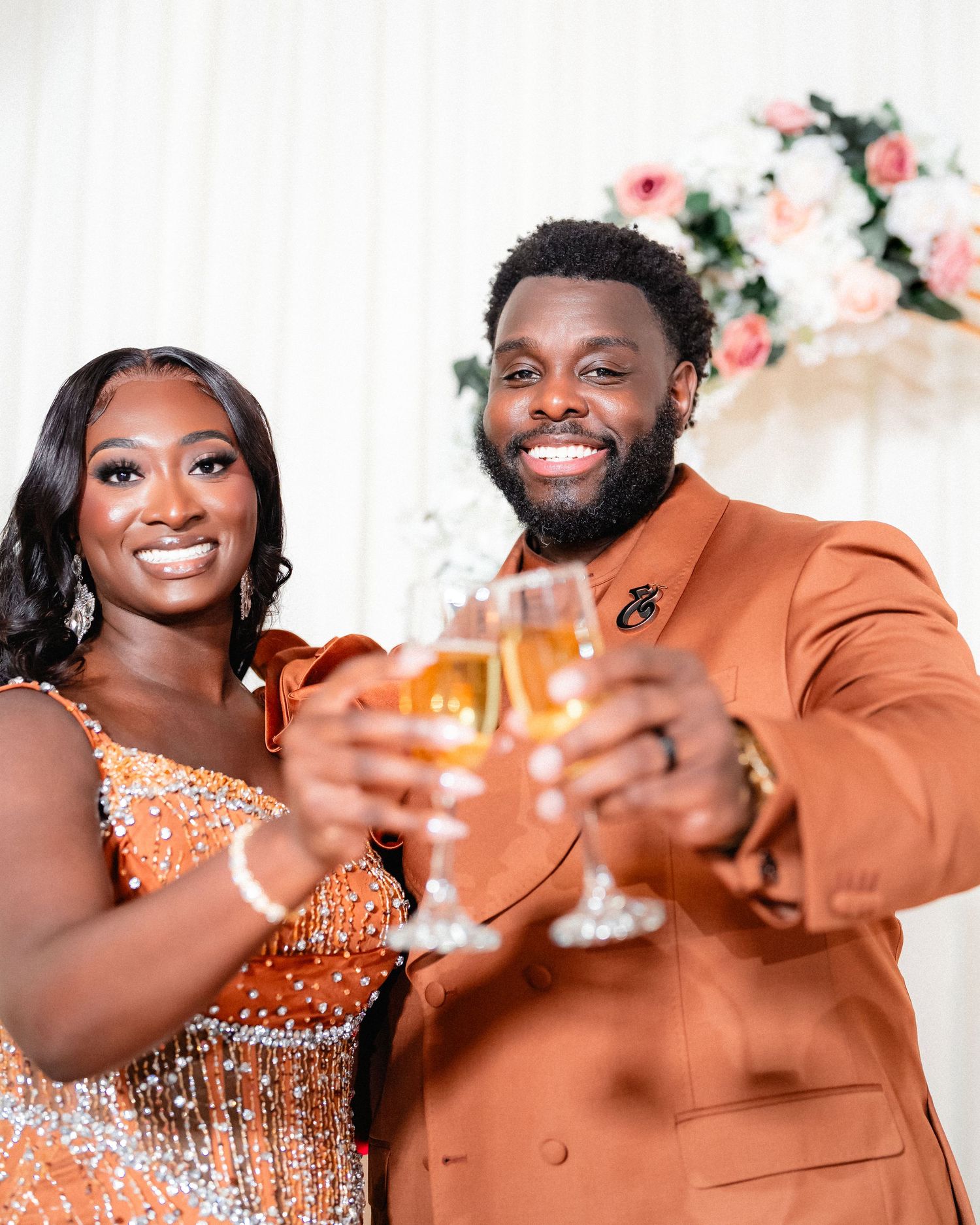 Wedding celebration with couple in formal attire toasting with champagne glasses against floral backdrop.